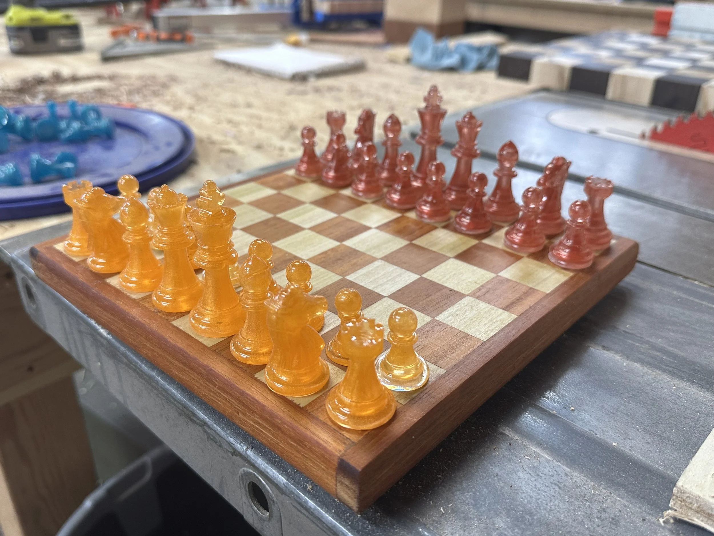 A chessboard with orange and red chess pieces set up for a game, on a wooden surface with tools and materials in the background.