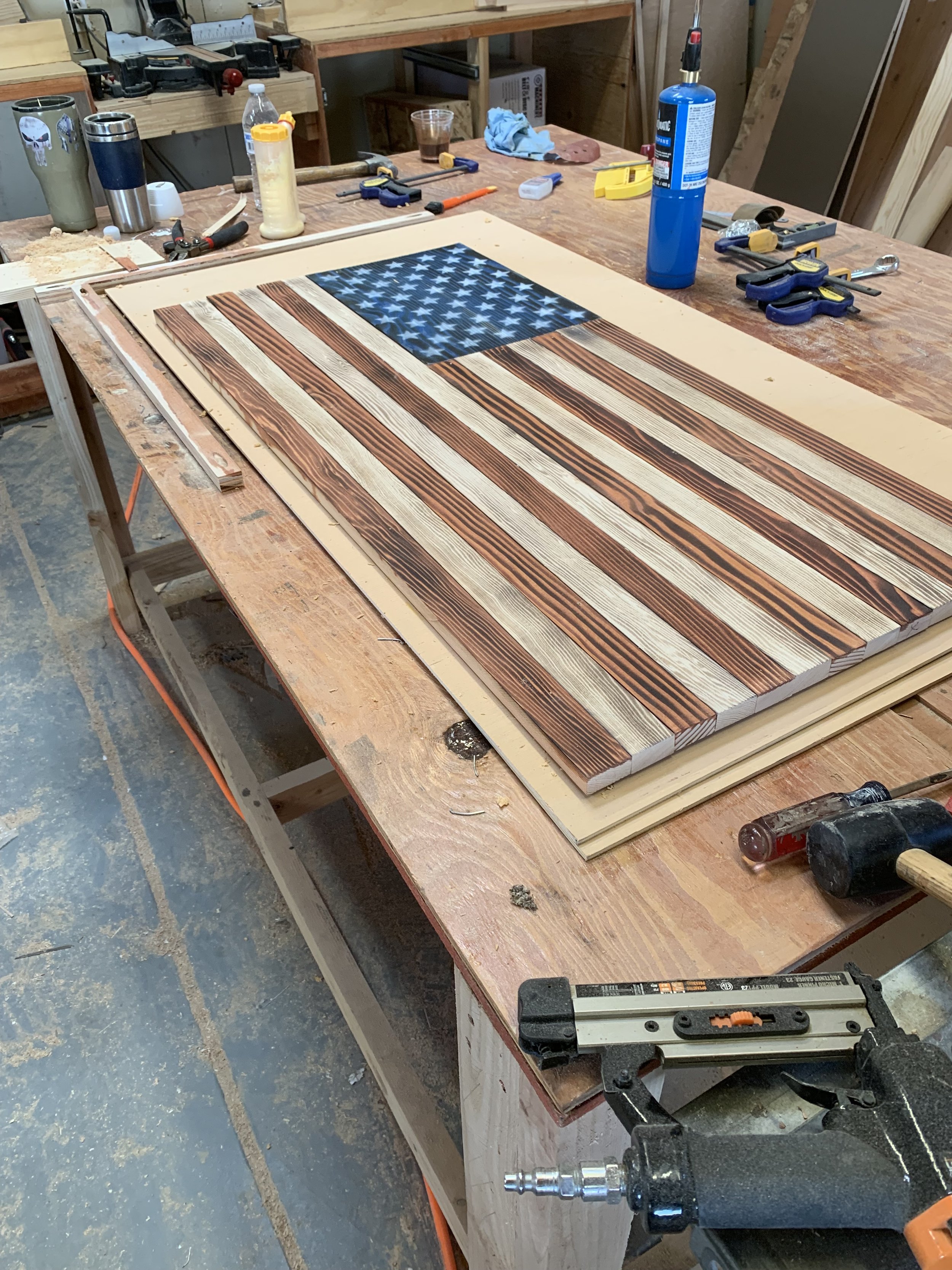 A woodworking workshop with a wooden table displaying a wooden American flag art piece under construction, various tools, and materials scattered around.