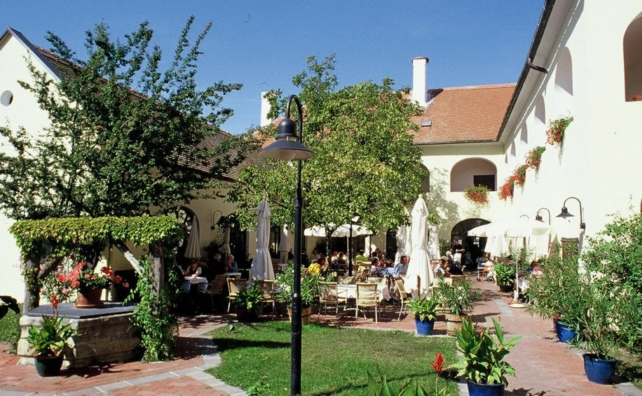 Außenansicht eines Restaurants oder Cafés mit Sitzplätzen im Freien, umgeben von Bäumen, Pflanzen in Töpfen und Sonnenschirmen.