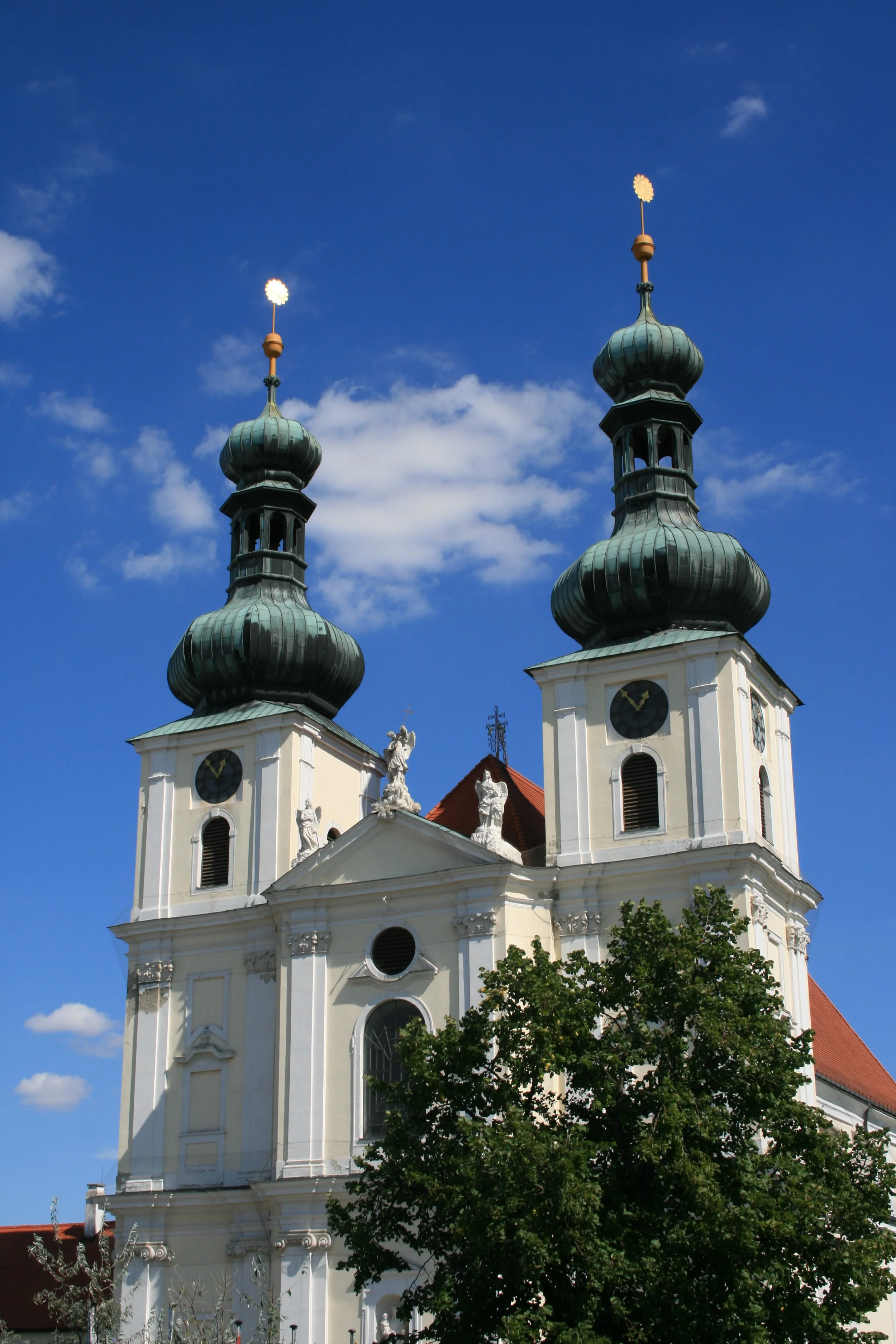 Barocke Kirche mit zwei Türmen und grünen Kuppeldächern vor blauem Himmel, mit Bäumen im Vordergrund.