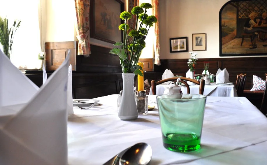 Ein festlich gedeckter Esstisch mit weißen Tischdecken, einer Vase mit grünen Blumen, Kerzen, Besteck, Gläsern und gefalteten Stoffservietten in einem Restaurant oder Café.