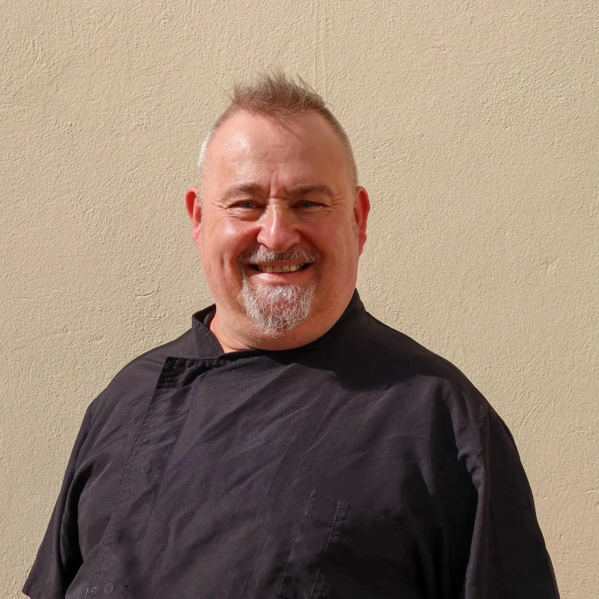 Ein Mann trägt eine schwarze Kochjacke und lächelt vor einem beigequerält.