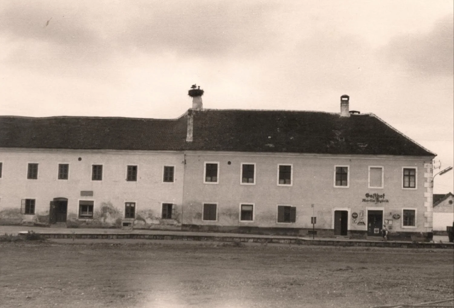 Ein altes, zweistöckiges Gebäude mit mehreren Fenstern und zwei Kaminen, eines mit einem Vogelnest. Das Gebäude hat eine Beschriftung, die 'Gasthof' und 'Reitstall' enthält, und befindet sich an einem Bahnhof mit Gleisen im Vordergrund.