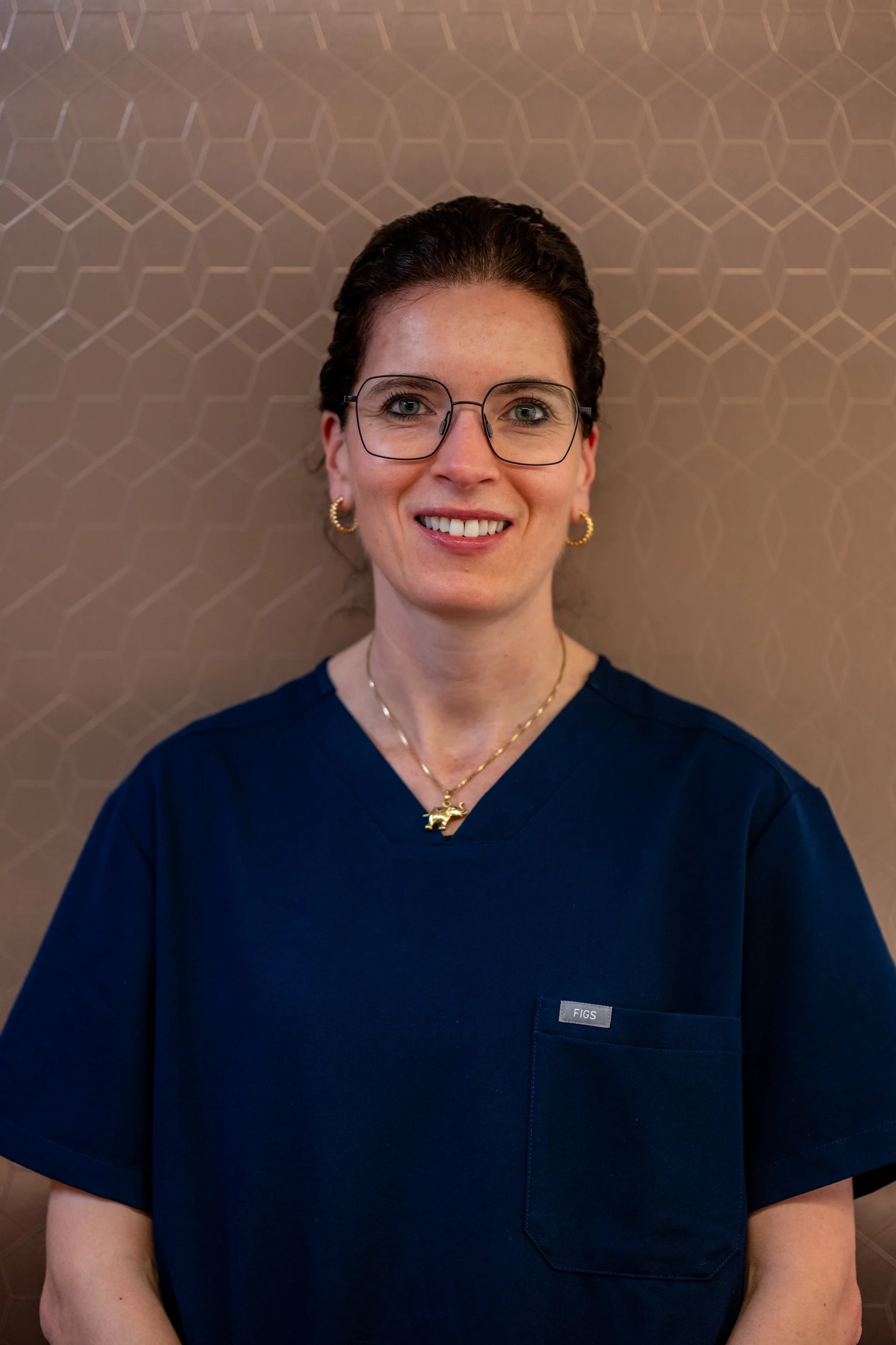 Eine Frau trägt eine dunkle medizinische Uniform, hat braune, zu einem Zopf zusammengebundene Haare, trägt Brille und goldene Schmuckstücke, steht vor einer braunen Wand mit geometrischem Muster.