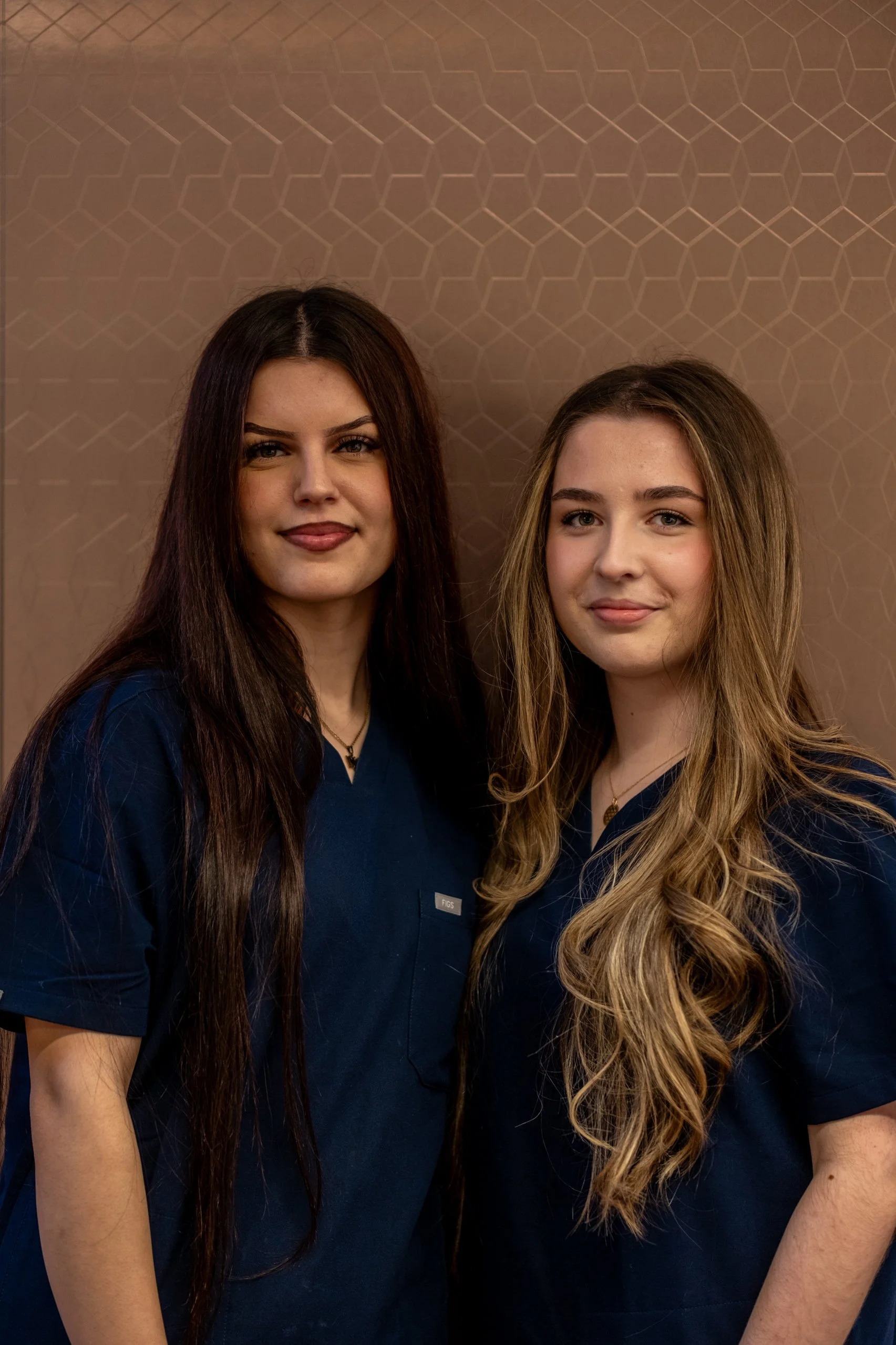 Zwei junge Frauen in blauen Arbeitskleidung stehen vor einer gepunkteten, braunen Wand.