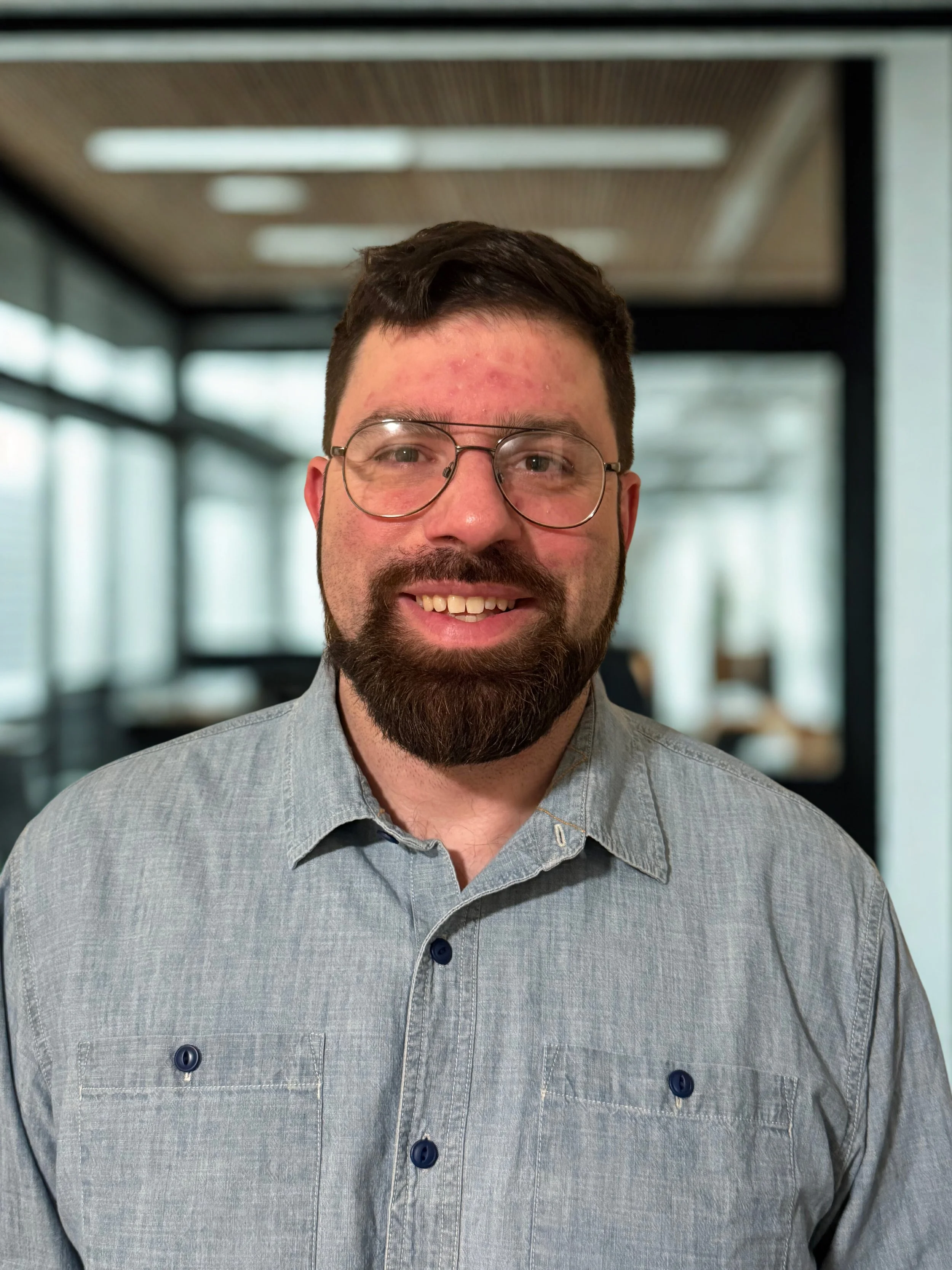 A smiling man with glasses and a beard, wearing a gray button-up shirt, standing in an office with large windows and modern interior design.