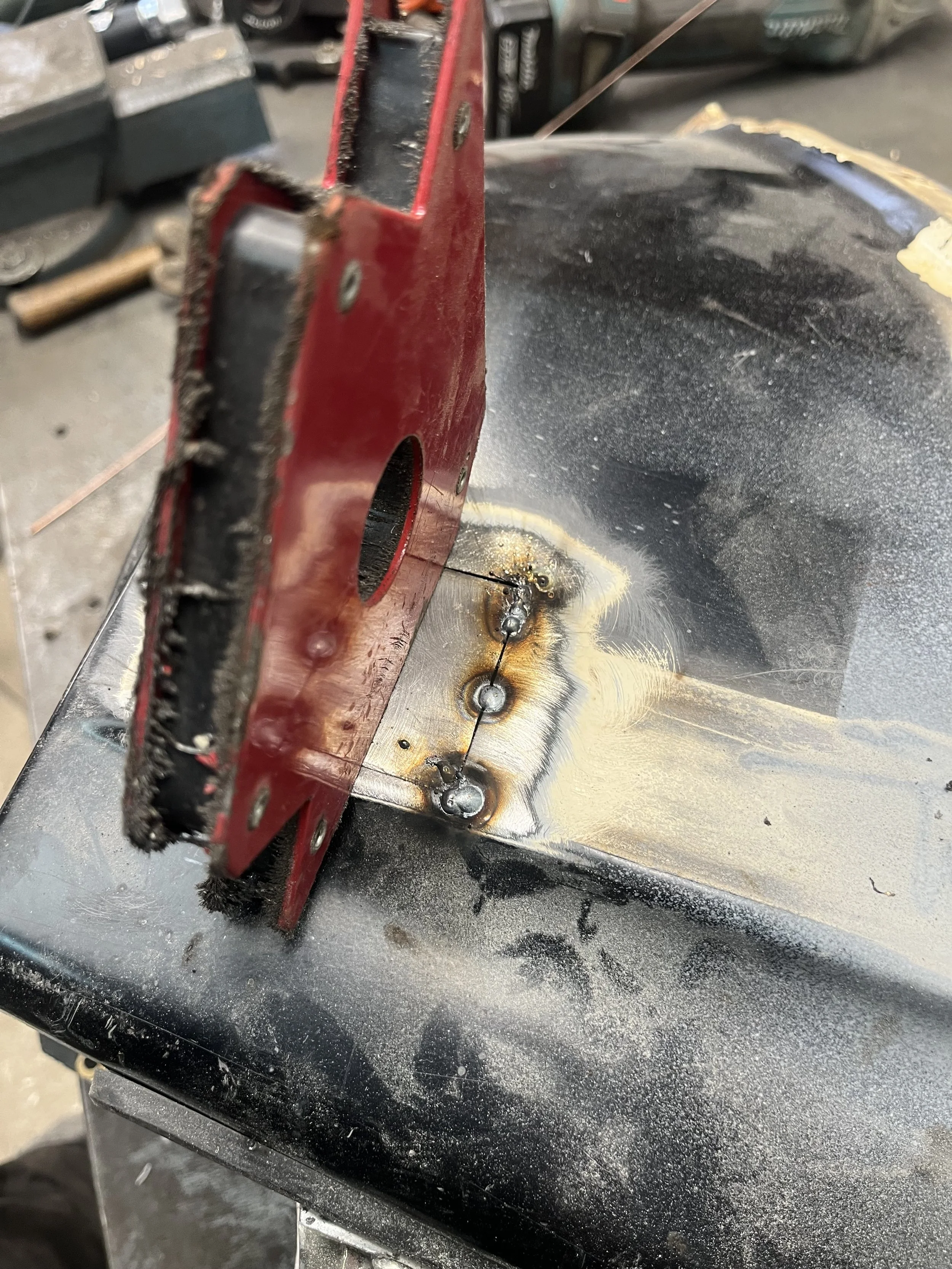 Close-up van een metalen werkstuk met twee gelaste verbindingen, gedeeltelijk bedekt door een rode lasklem, op een zwart werkoppervlak met zichtbare lasmarkeringen en asresten.