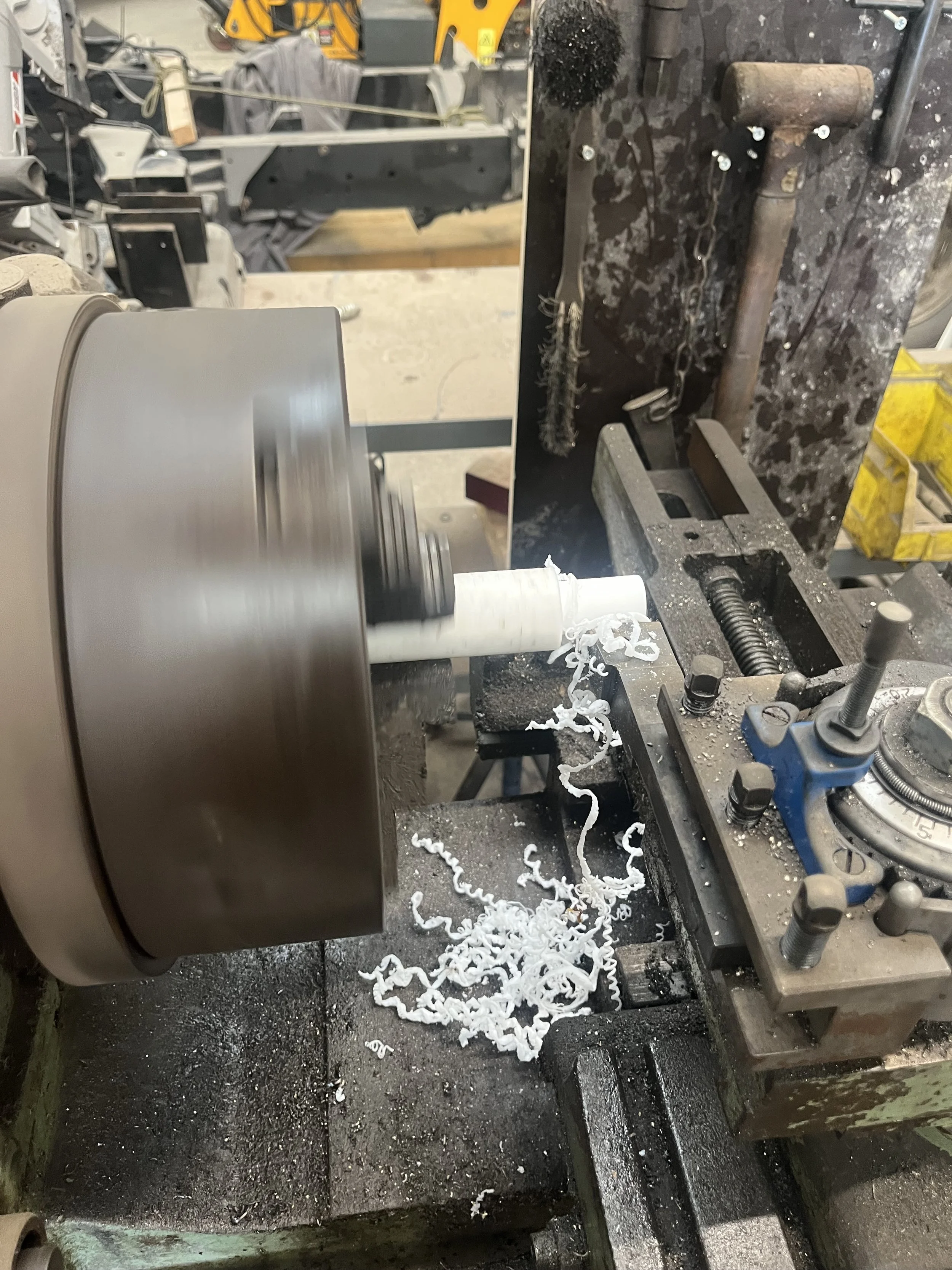 Een metaaldraaibank is een witte kunststofbuis aan het bewerken, met kunststofkrullen verspreid rondom en een metalen werkstuk gemonteerd op de draaibank.