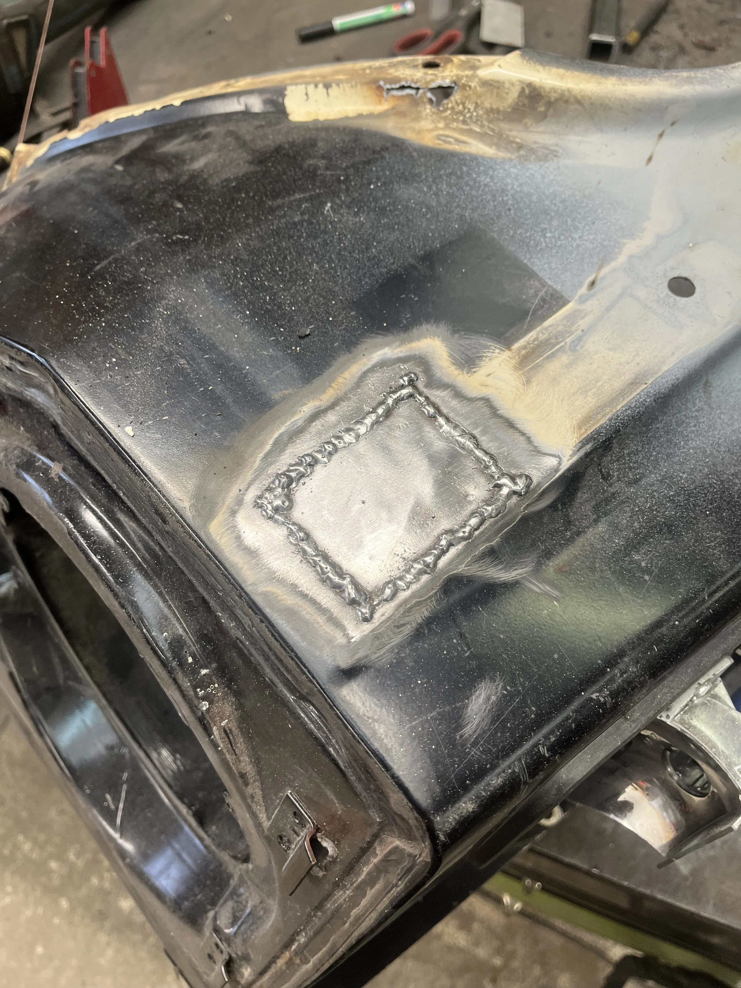 Een close-up van een metalen auto-onderdeel, vermoedelijk een motorkap of spatbord, met een vierkante patch die gelast of hersteld werd.