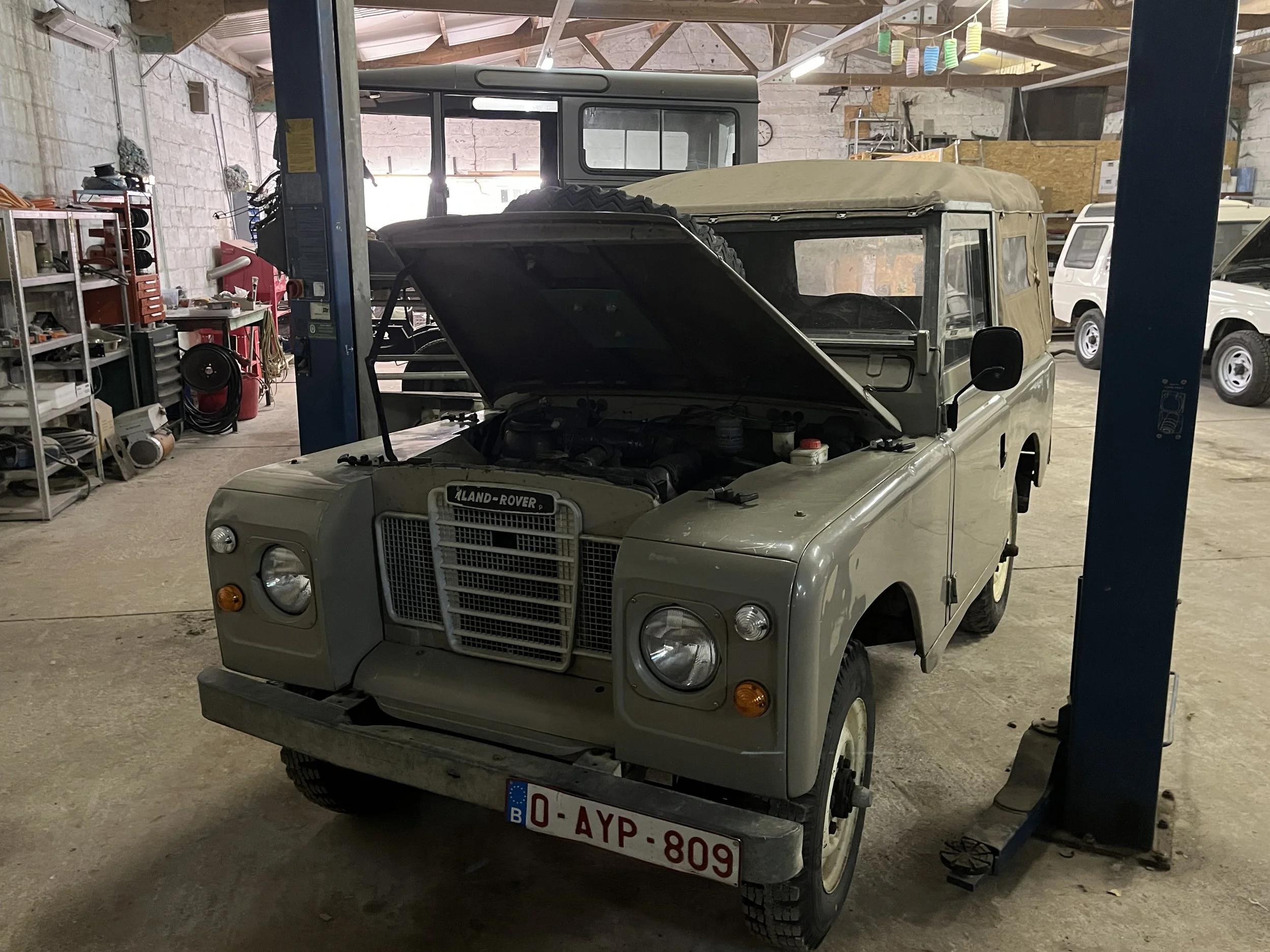 Een klassieke beige Land Rover met open motorkap in een werkplaats of garage. Links zijn verschillende gereedschappen en materialen zichtbaar op rekken, met op de achtergrond enkele auto’s en een heftruck.