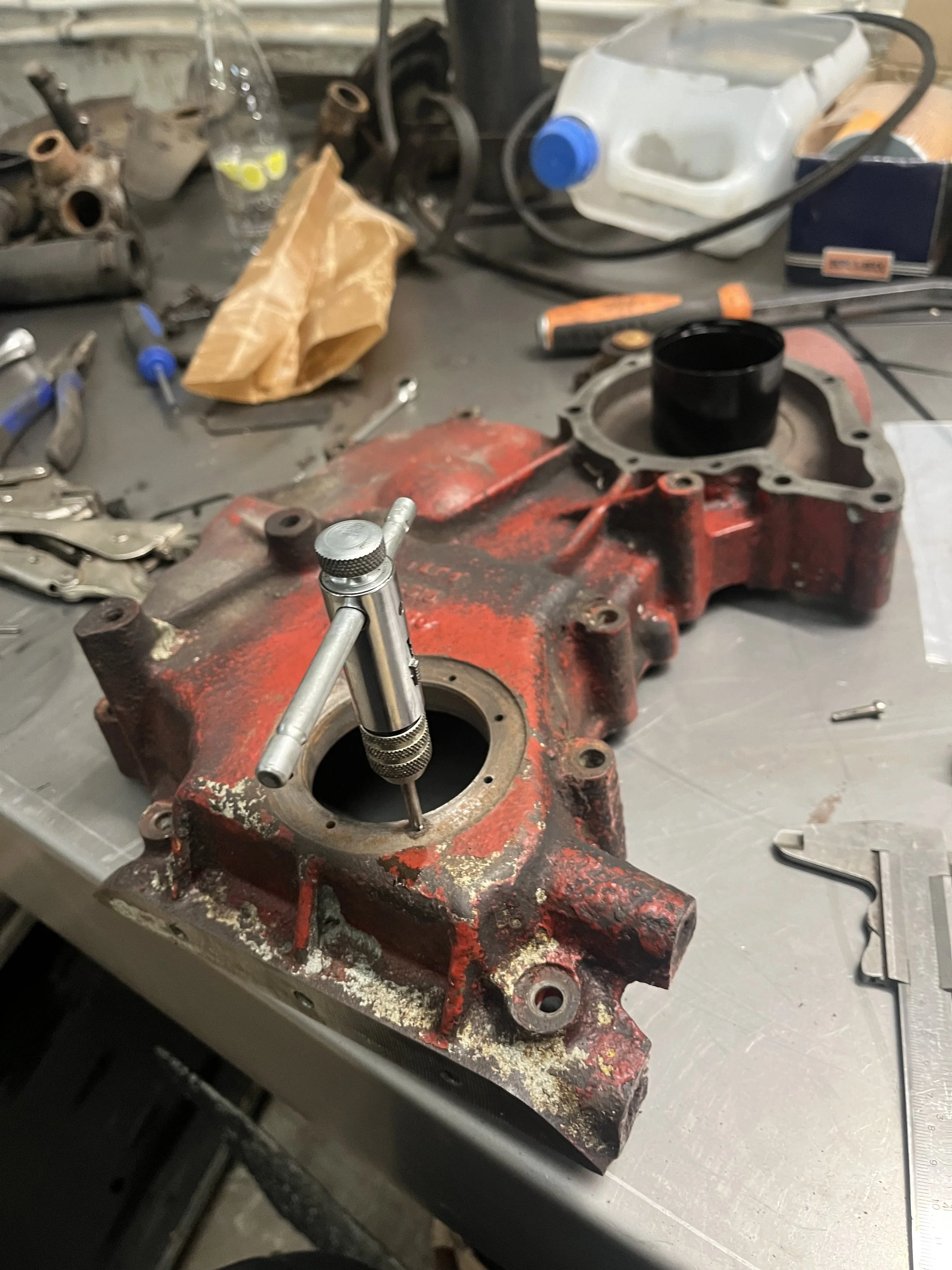 Een werkbank voor motorreparatie met een gedemonteerd rood motorblok, een cilindermeter die de cilinder controleert, en verschillende gereedschappen en benodigdheden verspreid op de tafel.