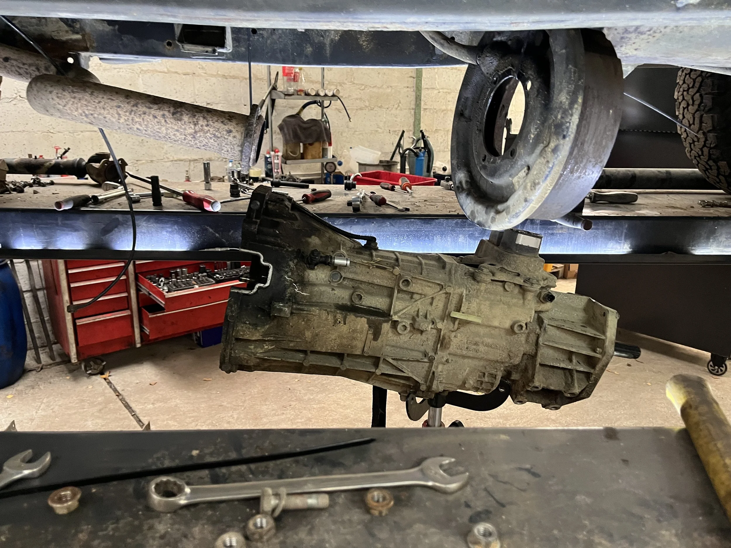 Automotive transmissie gemonteerd onder een voertuig op een werkbank in een garage, met gereedschap en apparatuur verspreid rondom.