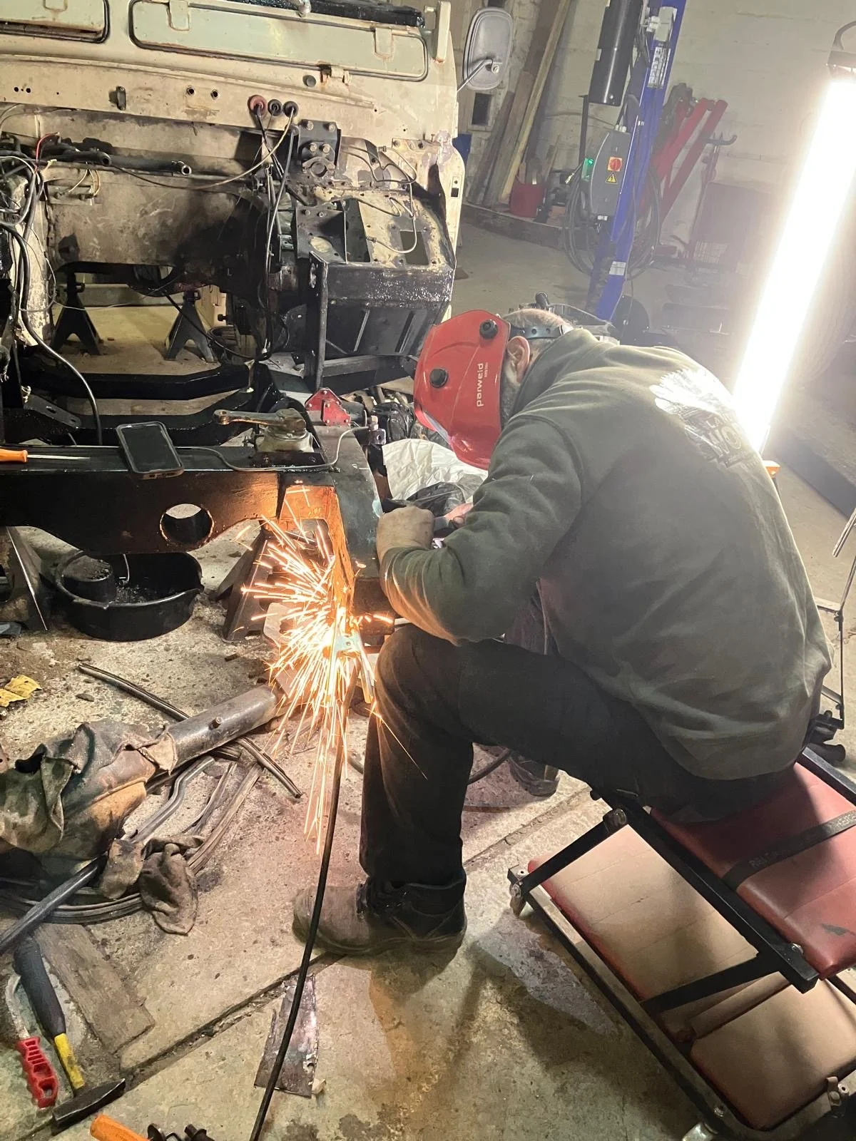 Een persoon met een rode lashelm en een camouflagejas is metaal aan het lassen in een werkplaats, terwijl hij op een klein bankje zit en vonken rondvliegen.