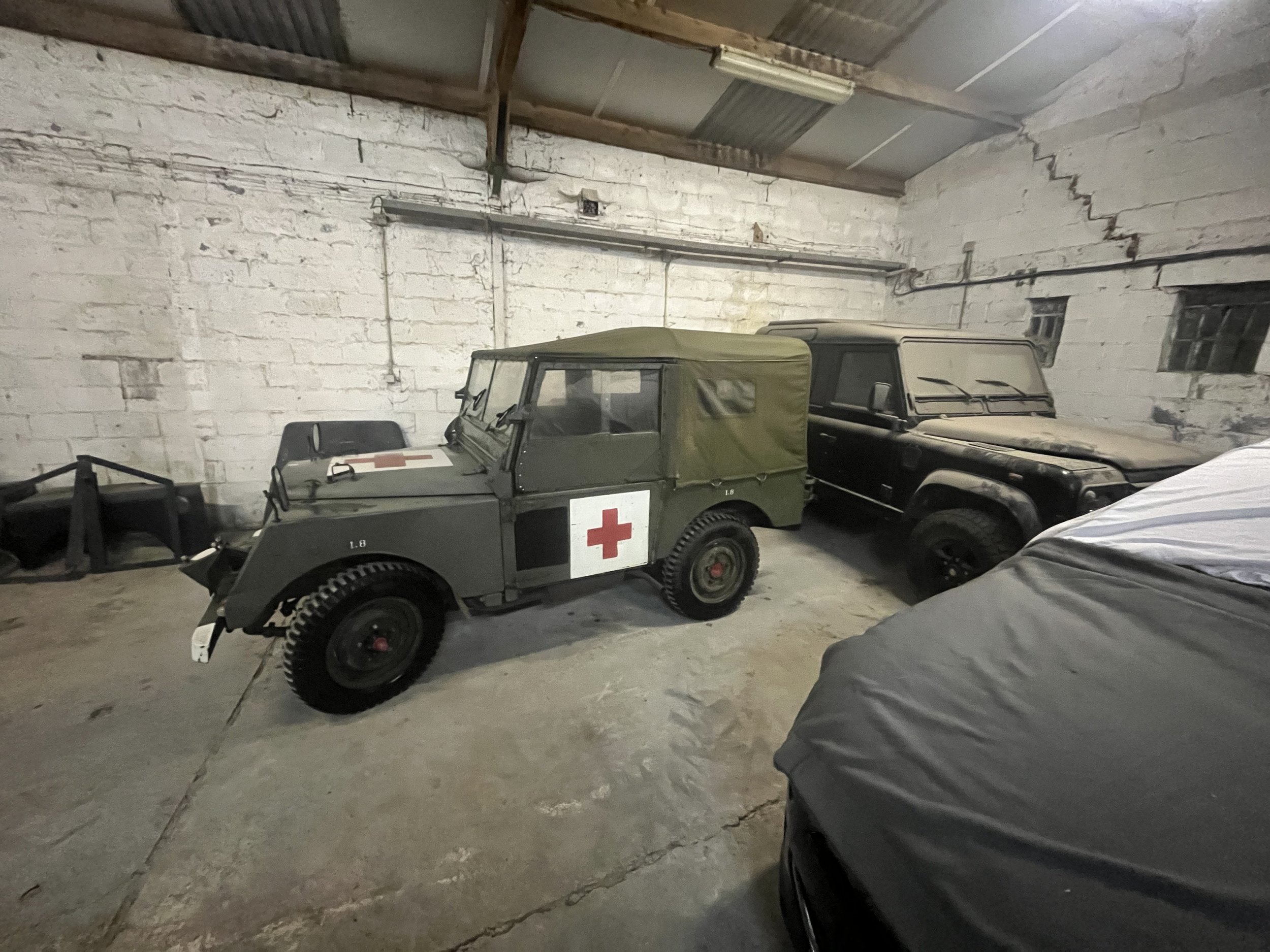 Twee vintage militaire voertuigen, waarvan één met een roodkruissymbool, geparkeerd in een betonnen garage met een ruwe bakstenen muur en een plafond van golfplaat.