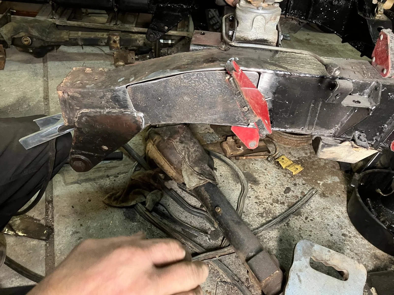 Close-up van het chassis en de ophangingscomponenten van een voertuig tijdens herstelling, met roest en vuil op de metalen onderdelen en een hand die aan de montage werkt in een garage of werkplaats.