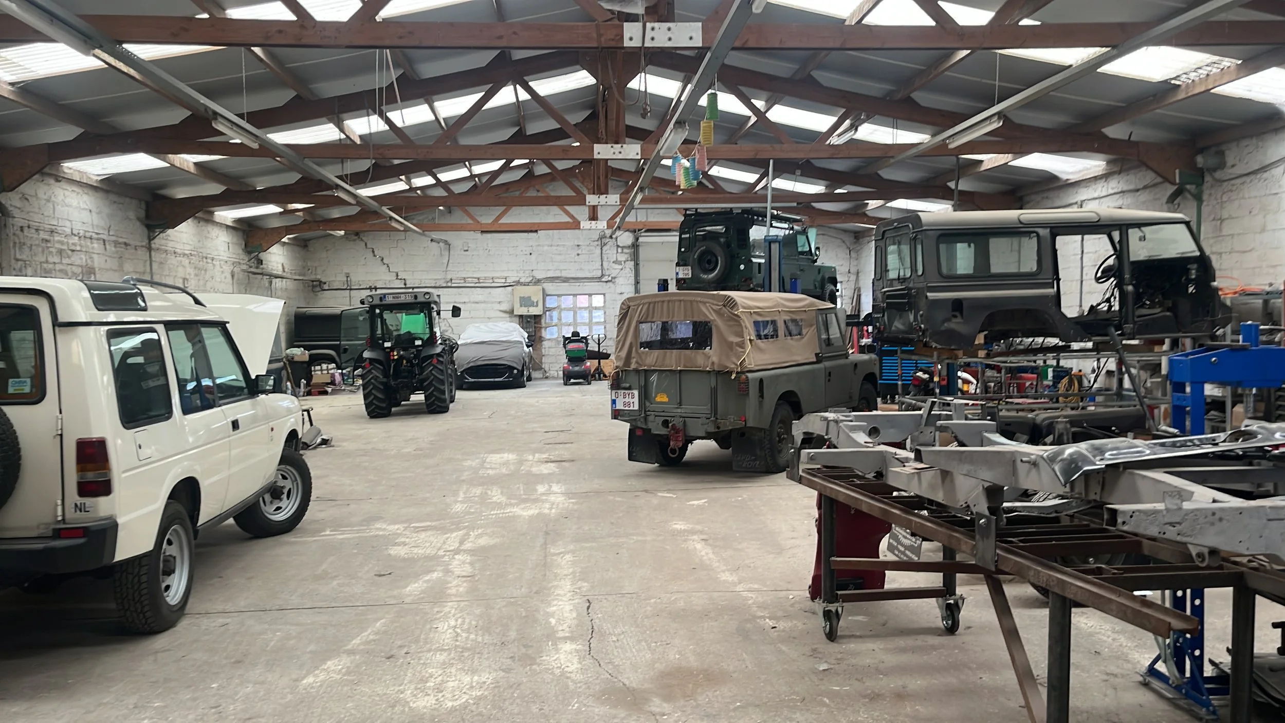 Interieur van een voertuigherstelgarage met meerdere voertuigen: links een witte bestelwagen, in het midden een militair voertuig en rechts een zwarte terreinwagen die op een hefbrug staat, met gereedschap en materiaal verspreid in de werkruimte.