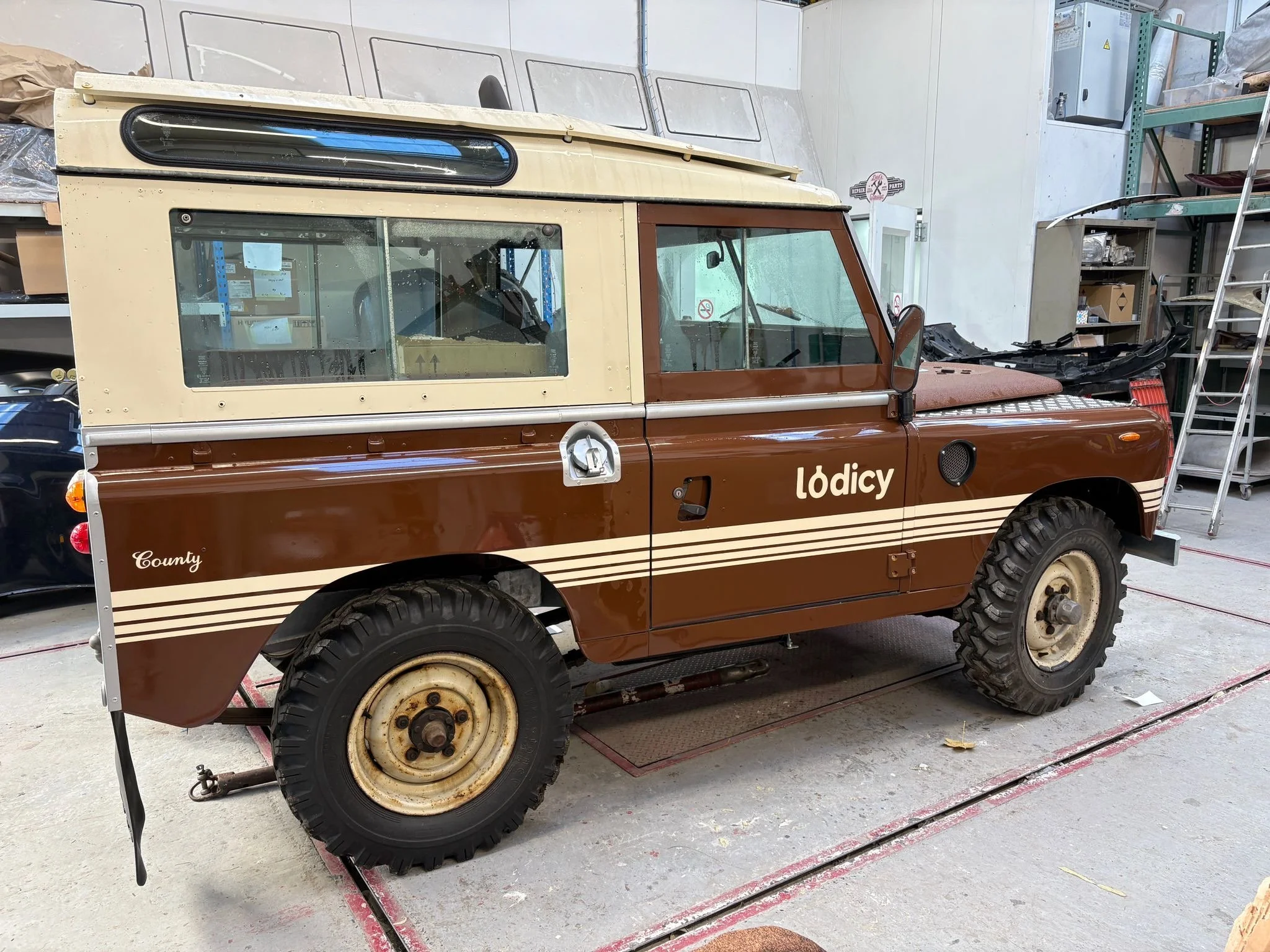 Een vintage bruin-crèmekleurig terreinvoertuig met het woord ‘Lodicy’ op de zijkant, geparkeerd in een werkplaats of opslagruimte.