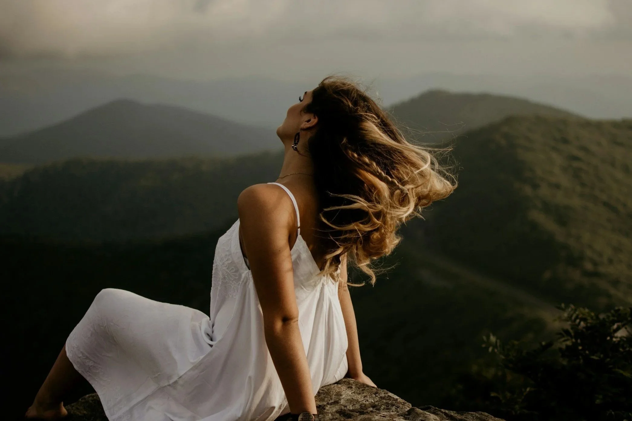 A woman with long wavy hair wearing a white dress, sitting on a large rock overlooking mountains and cloudy sky with wind blowing her hair.