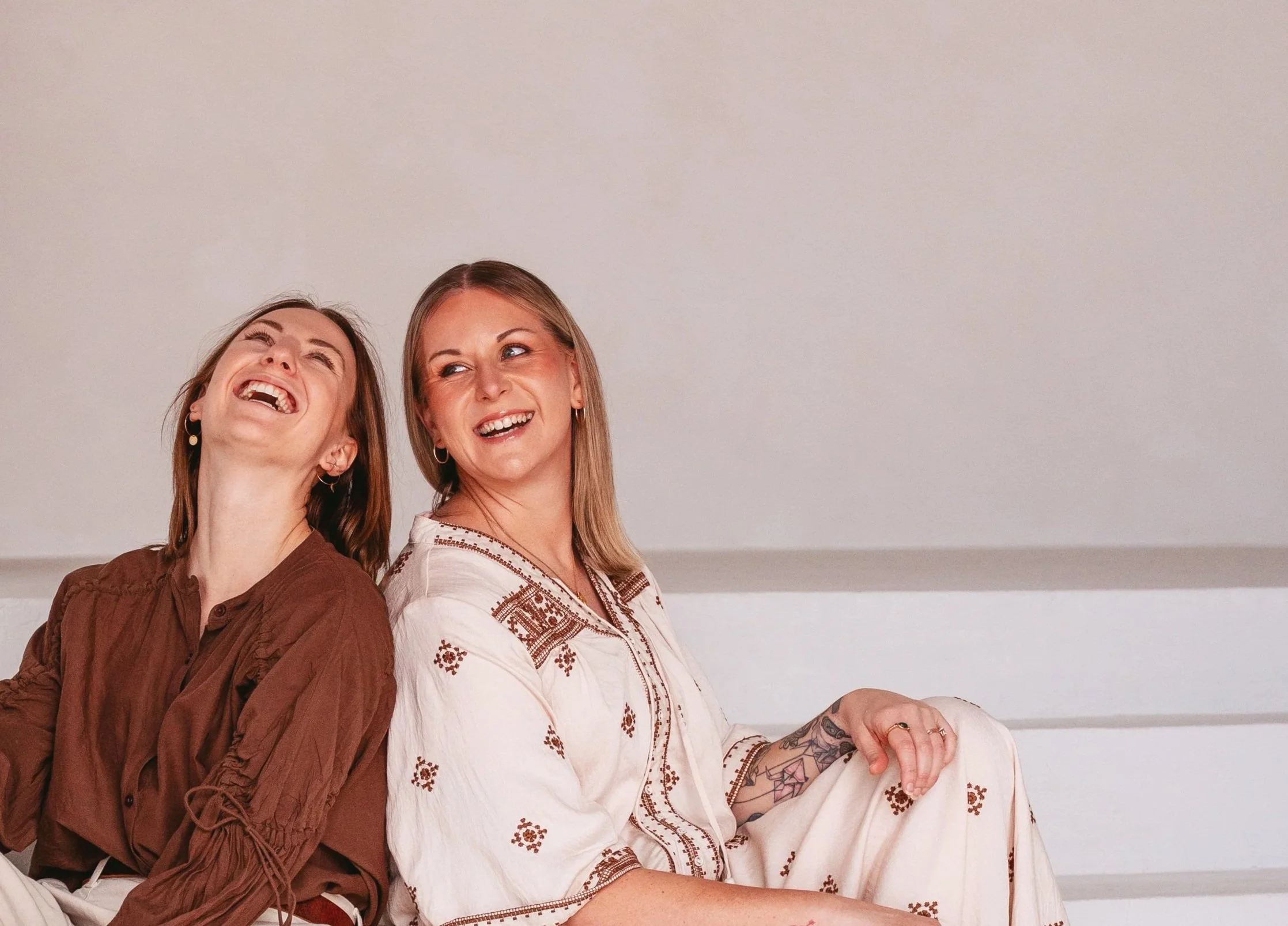 Two women are sitting and smiling, with one looking upwards and the other looking sideways. They are against a plain light-colored wall.