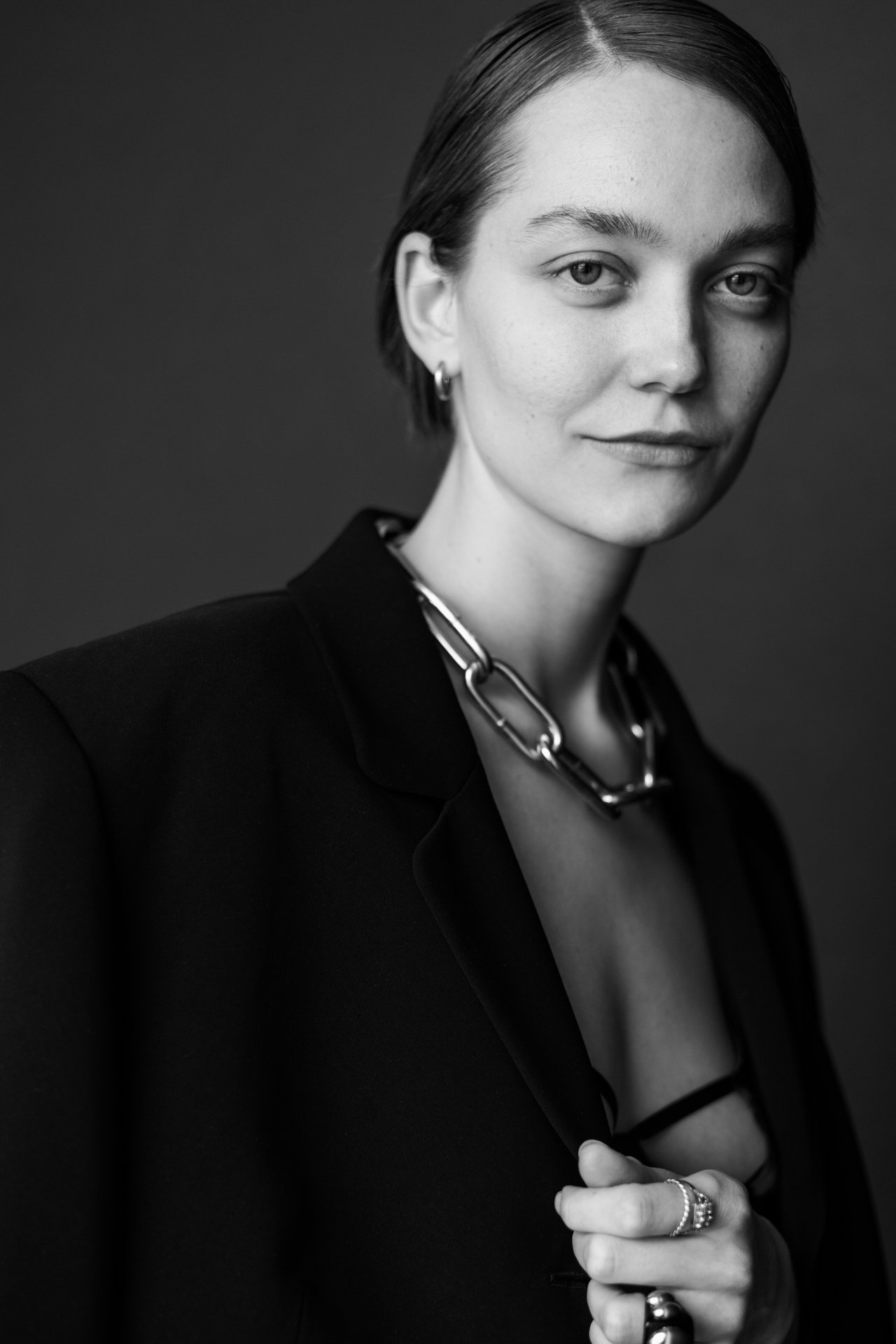 Schwarz-Weiß-Fotografie einer jungen Frau mit kurzen, glatten Haaren, die einen schwarzen Blazer trägt, viele silberne Schmuckstücke, darunter eine auffällige Kette und Ringe, und die einen entspannten Gesichtsausdruck hat.