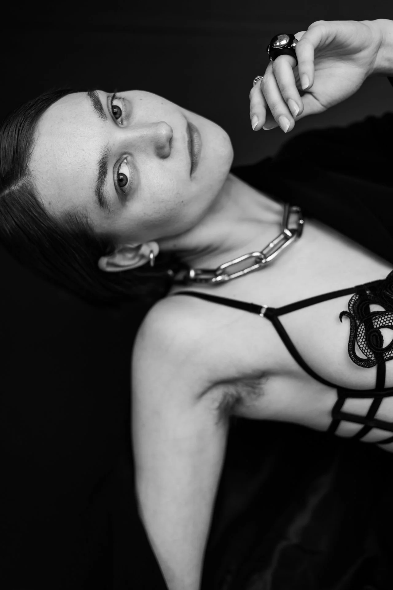 Schwarze und weiße Nahaufnahme eines jungen Frauen mit dunklen Haaren, Augenkontakt mit der Kamera, trägt eine große Kette, schwarze Spitze Kleidung, und mehrere Ringe, vor einem dunklen Hintergrund.