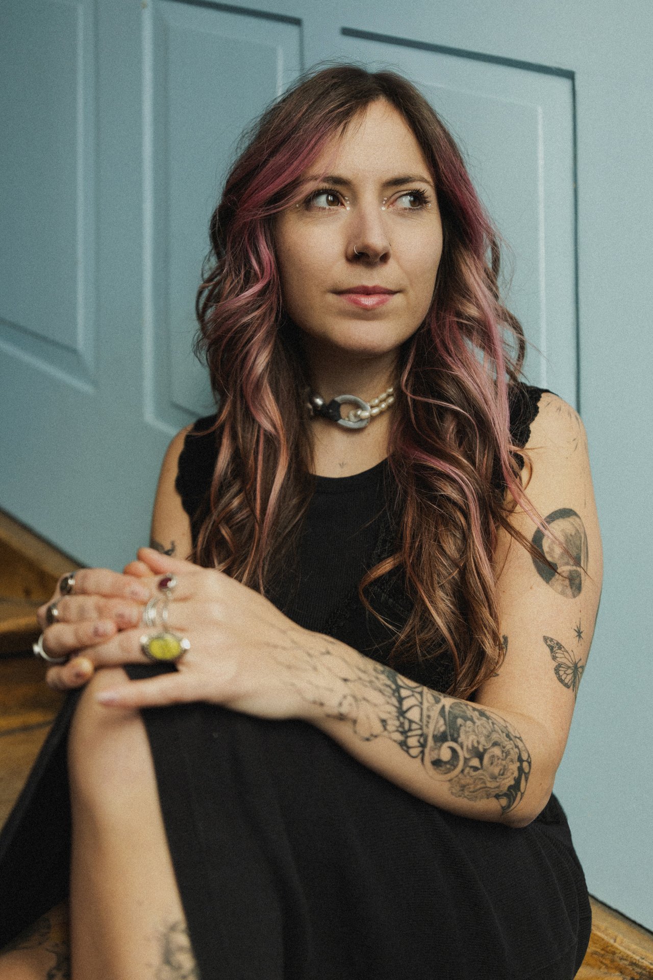 Eine junge Frau mit langem, welligem, pink-gefärbtem Haar sitzt vor einer blauen Wand und trägt mehrere Ringe, ein Halsband und Tattoos am Arm.