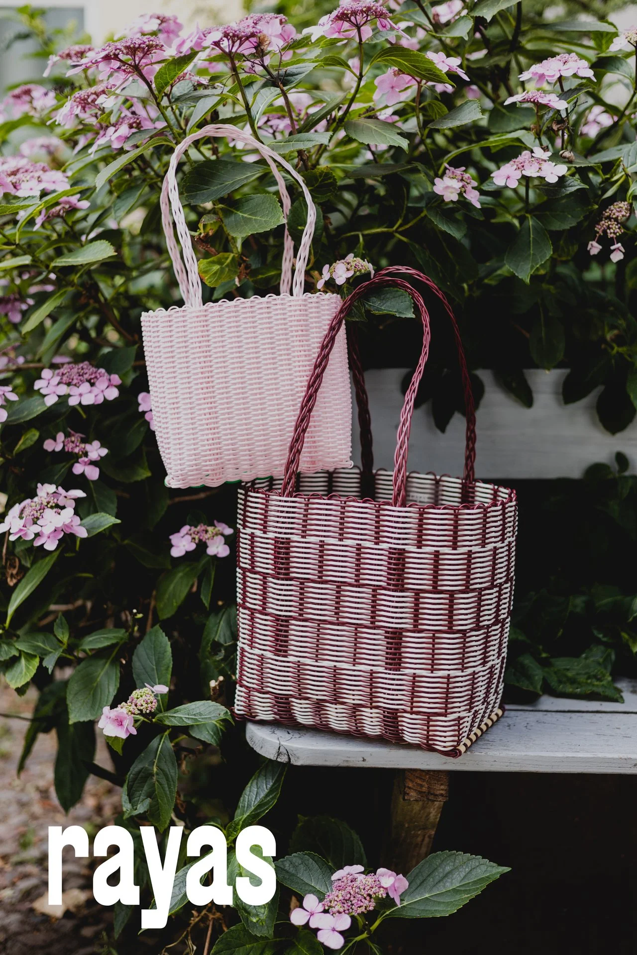 Zwei geflochtene Körbe, einer klein und einer groß, hängen an einer hibiskuspflanze mit pinken Blüten, vor einer weißen Holzbank. Der Text 'rays' ist im unteren linken Teil des Bildes teilweise sichtbar.