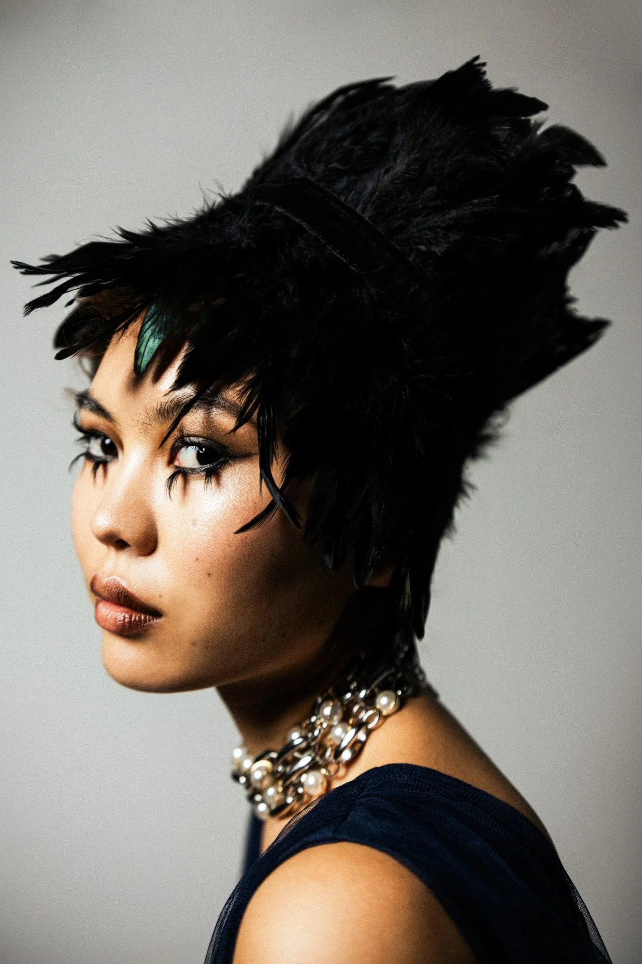 Nahaufnahme eines Models mit schwarzen Federn auf dem Kopf, dunklem Make-up und Perlenkette, seitliche Ansicht.