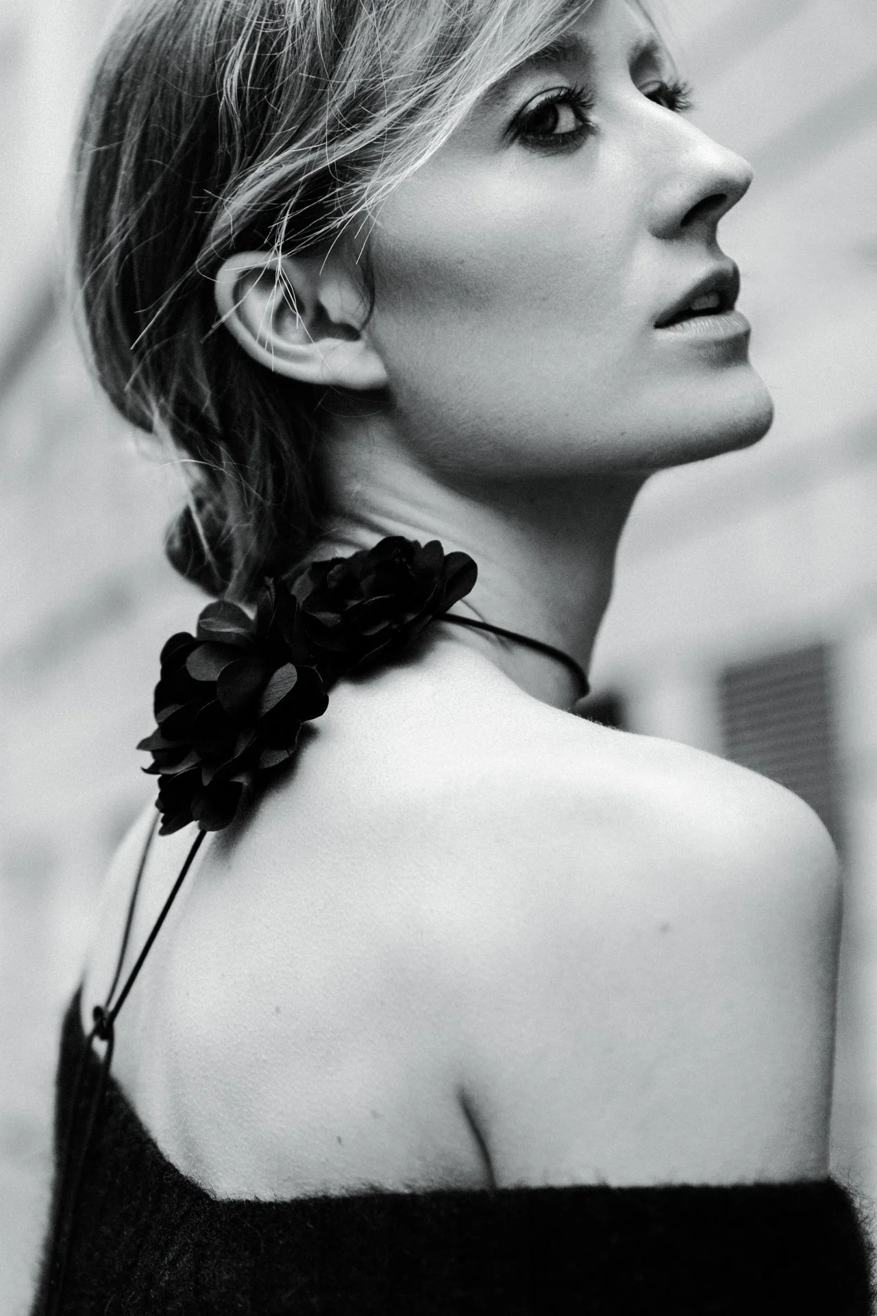 Schwarz-Weiß-Fotografie einer Frau mit kurzen Haaren, die nach rechts blickt, eine schwarze Blume auf der Schulter trägt, mit einem schwarzen Kleid, das auf der linken Seite sichtbar ist.
