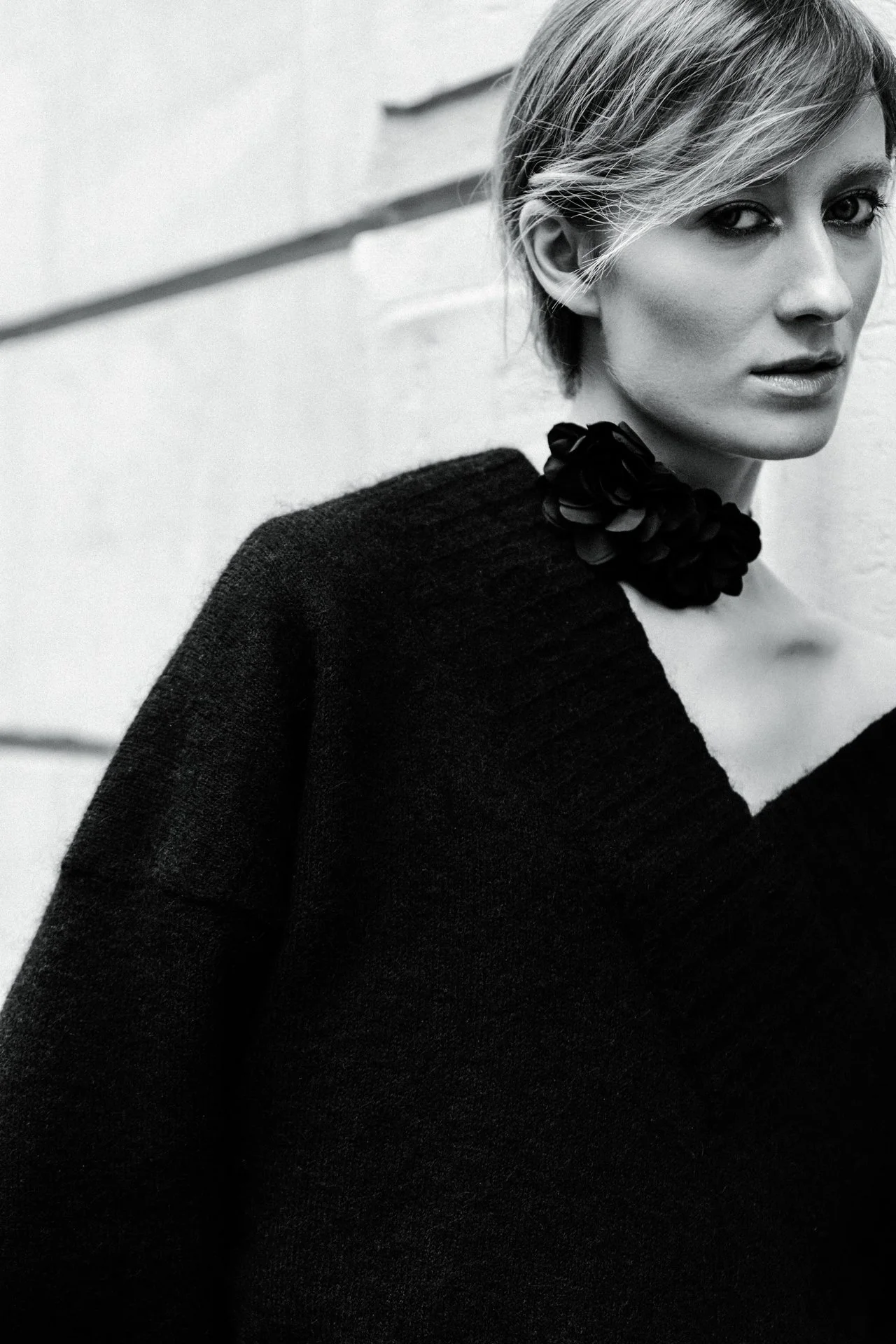 Schwarz-weiße Porträtaufnahme einer Frau mit kurzen, gestuften Haaren, die einen schwarzen Pullover trägt. Sie trägt einen schwarzen Blumenkragen oder -schmuck um den Hals und blickt direkt in die Kamera, während sie an einer Wand steht.