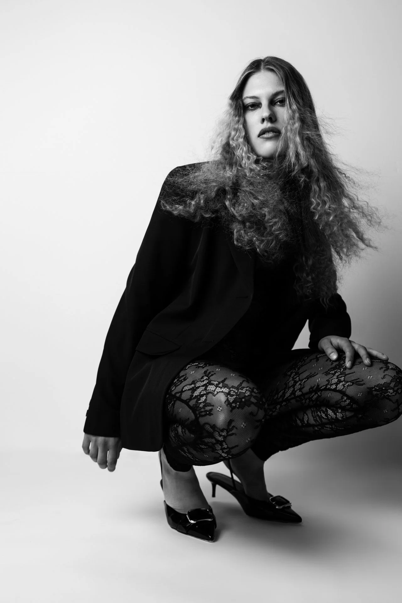 Schwarz-Weiß-Fotografie einer jungen Frau mit lockigem Haar, die eine schwarze Jacke trägt, gestehend auf einem Bein, mit hohen Schuhen und Netzhosen, vor hellem Hintergrund.