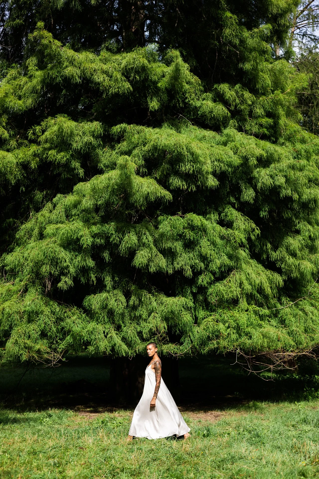 Eine junge Frau in einem weißen Kleid steht vor einem großen grünen Baum im Freien.