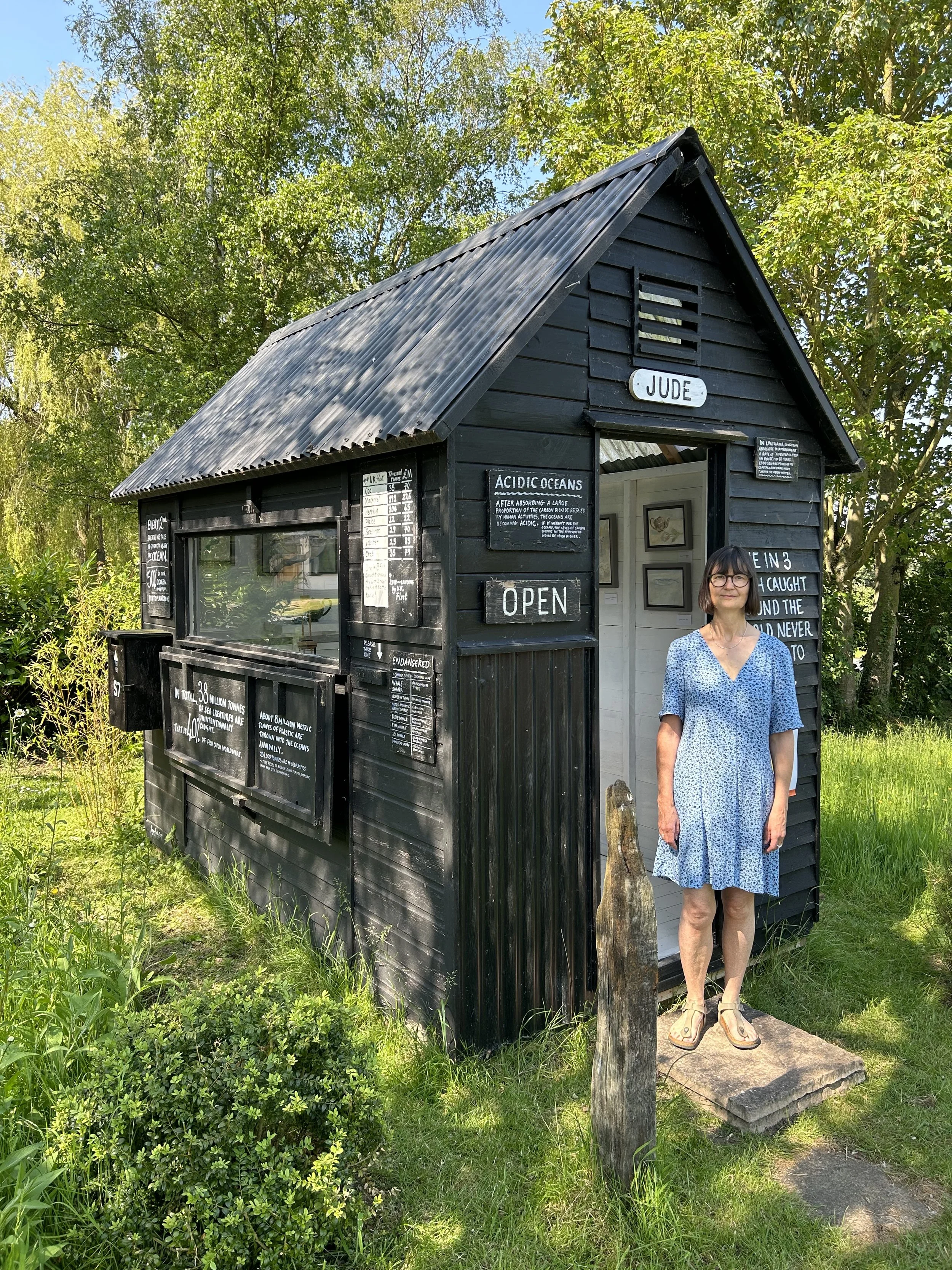 A woman standing outside a small black wooden house with signs about ocean acidity and conservation, surrounded by greenery and trees.