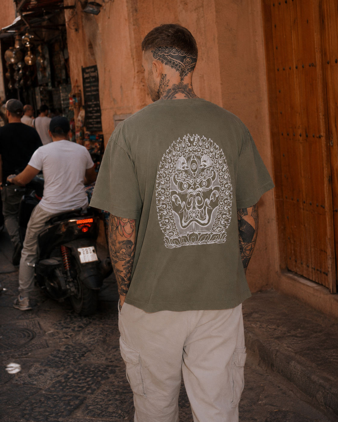 A man with tattoos on his arms and neck is walking down a street with a large tattoo of a fierce lion on the back of his green T-shirt.