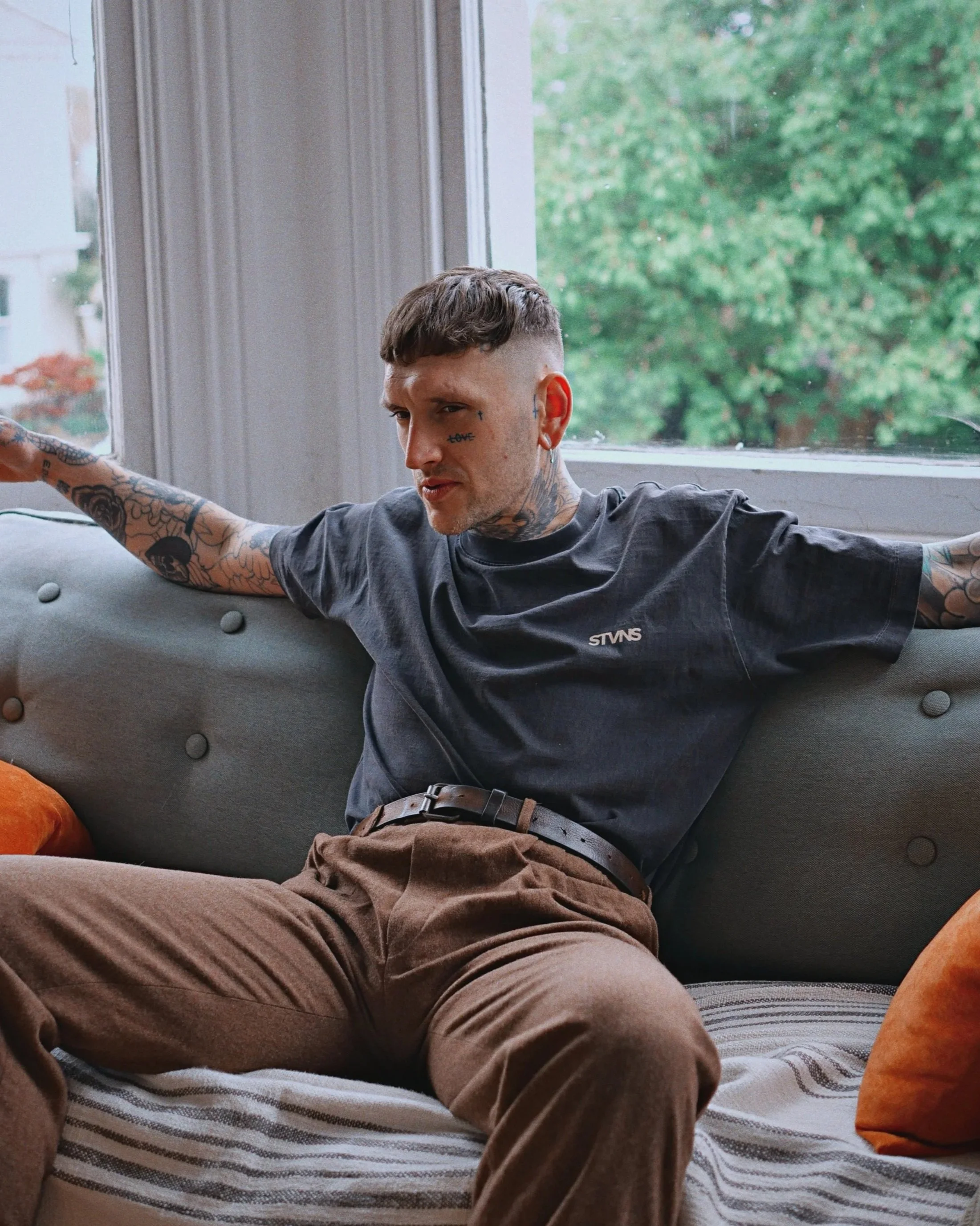A man with tattoos, short hair, and wearing a gray T-shirt sitting on a couch, with his arms extended along the back of the couch, in front of a large window with a view of green trees outside.