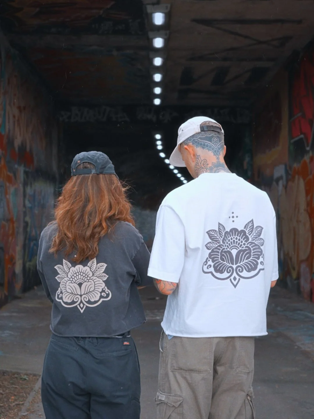 A man and a woman walking in an urban tunnel with graffiti on the walls, both wearing shirts with a similar stylized floral design on the back. The man has tattoos on his neck and wears a baseball cap backward, while the woman has long, wavy hair and