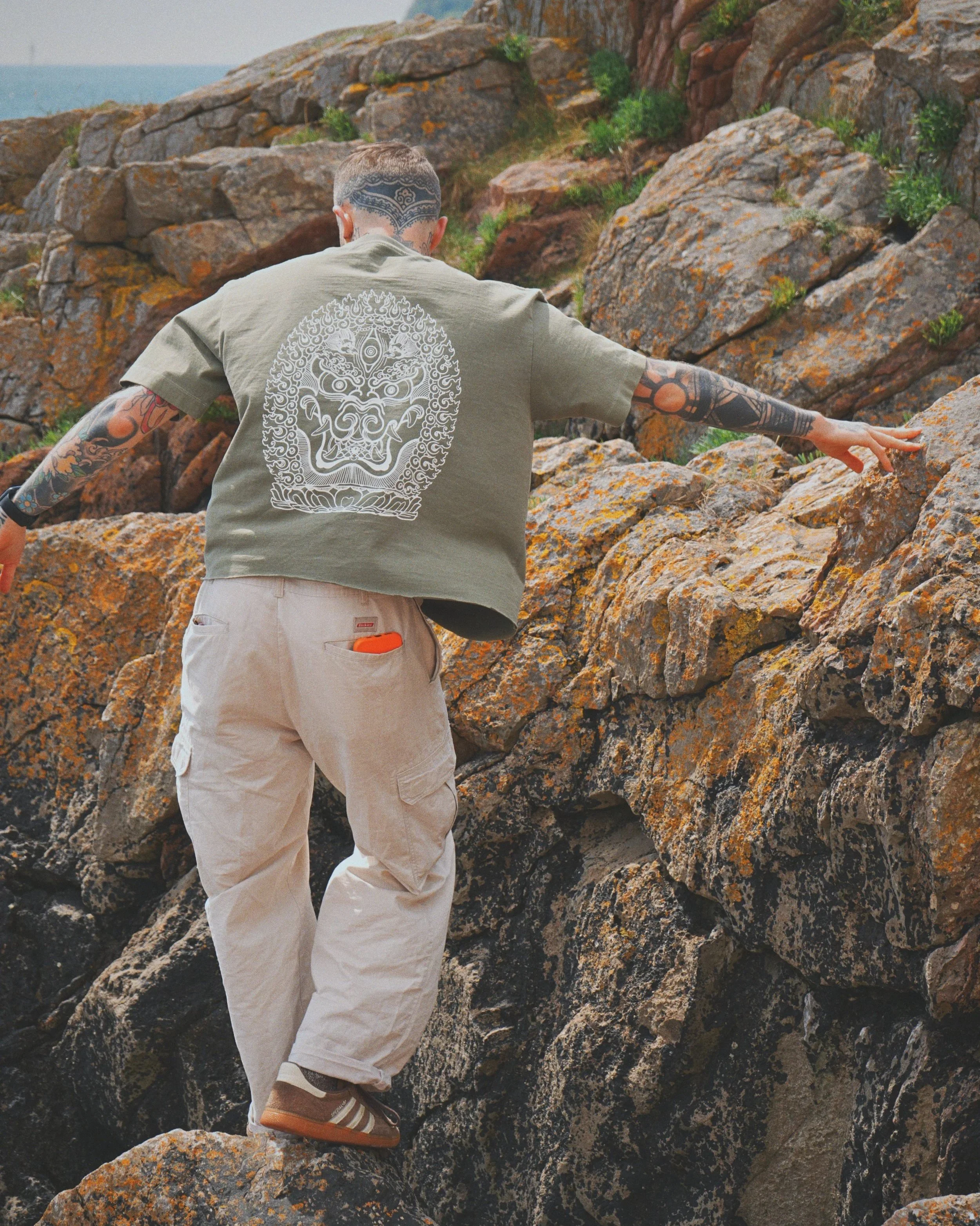 Man with tattoos climbing rocky terrain near water, wearing beige pants, brown sneakers, and a gray t-shirt with a detailed white graphic on the back.