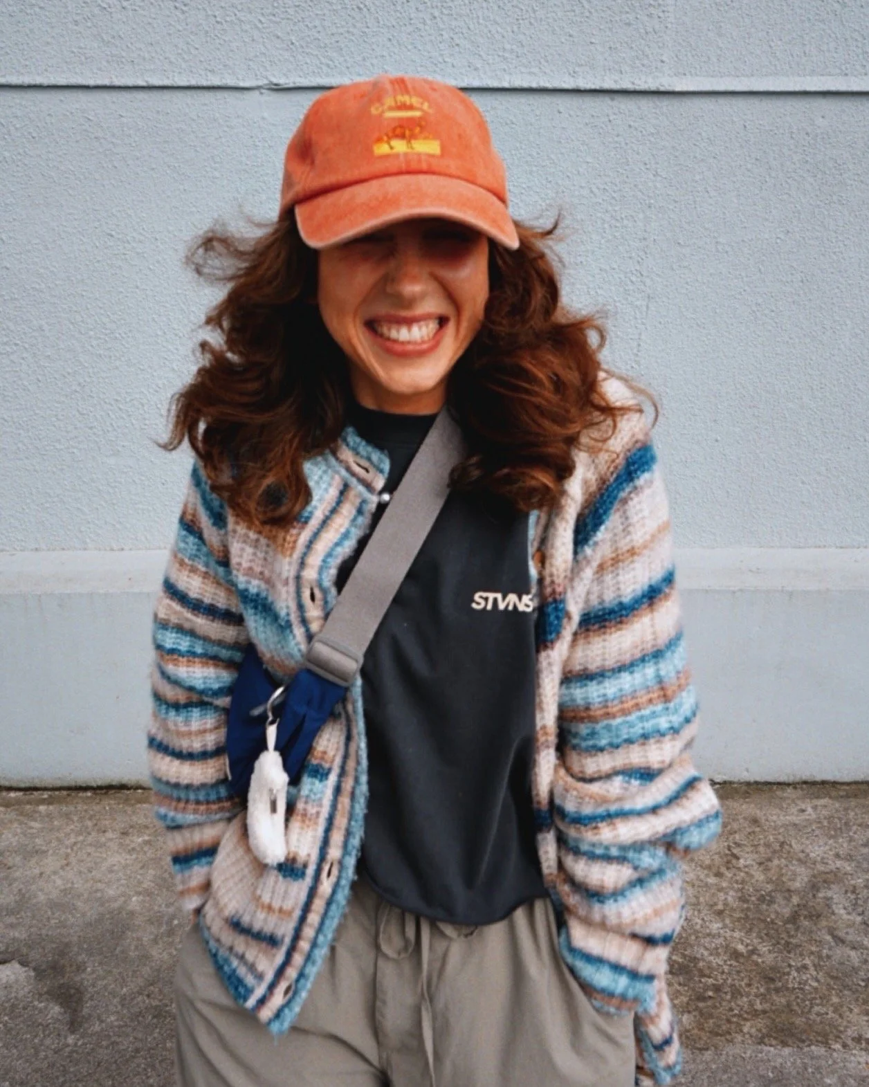 A woman with curly brown hair smiling, wearing a red cap, a multicolored striped cardigan, a black t-shirt, and beige pants, standing outdoors against a light gray wall.