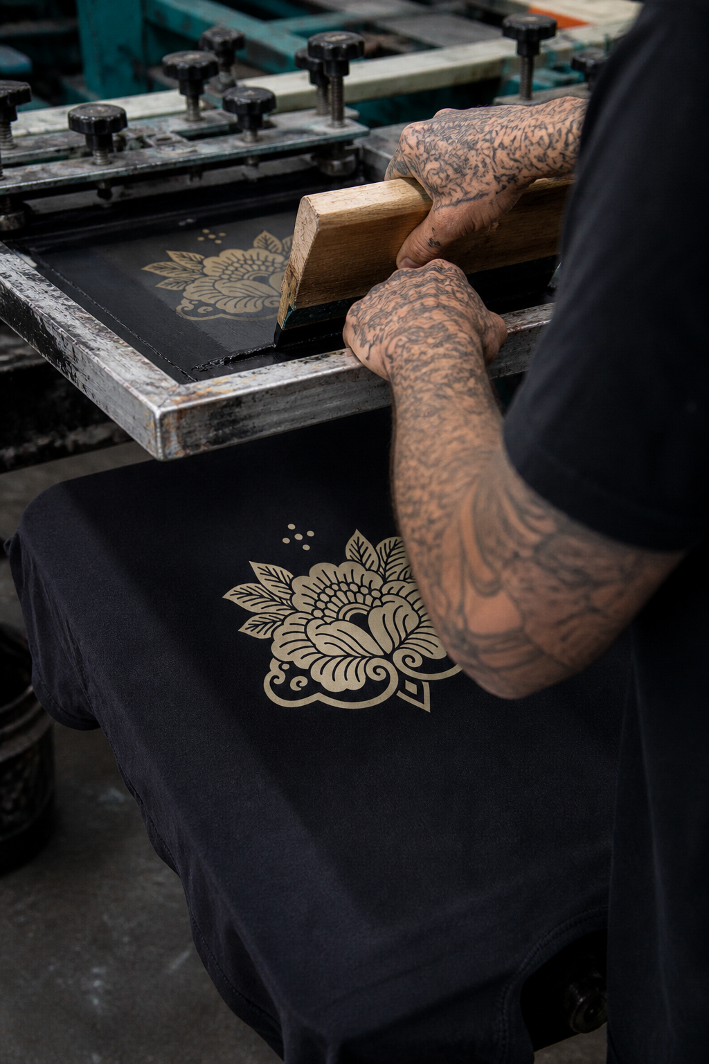 Person with tattooed arms working on printing a floral design on fabric using a screen printing frame.