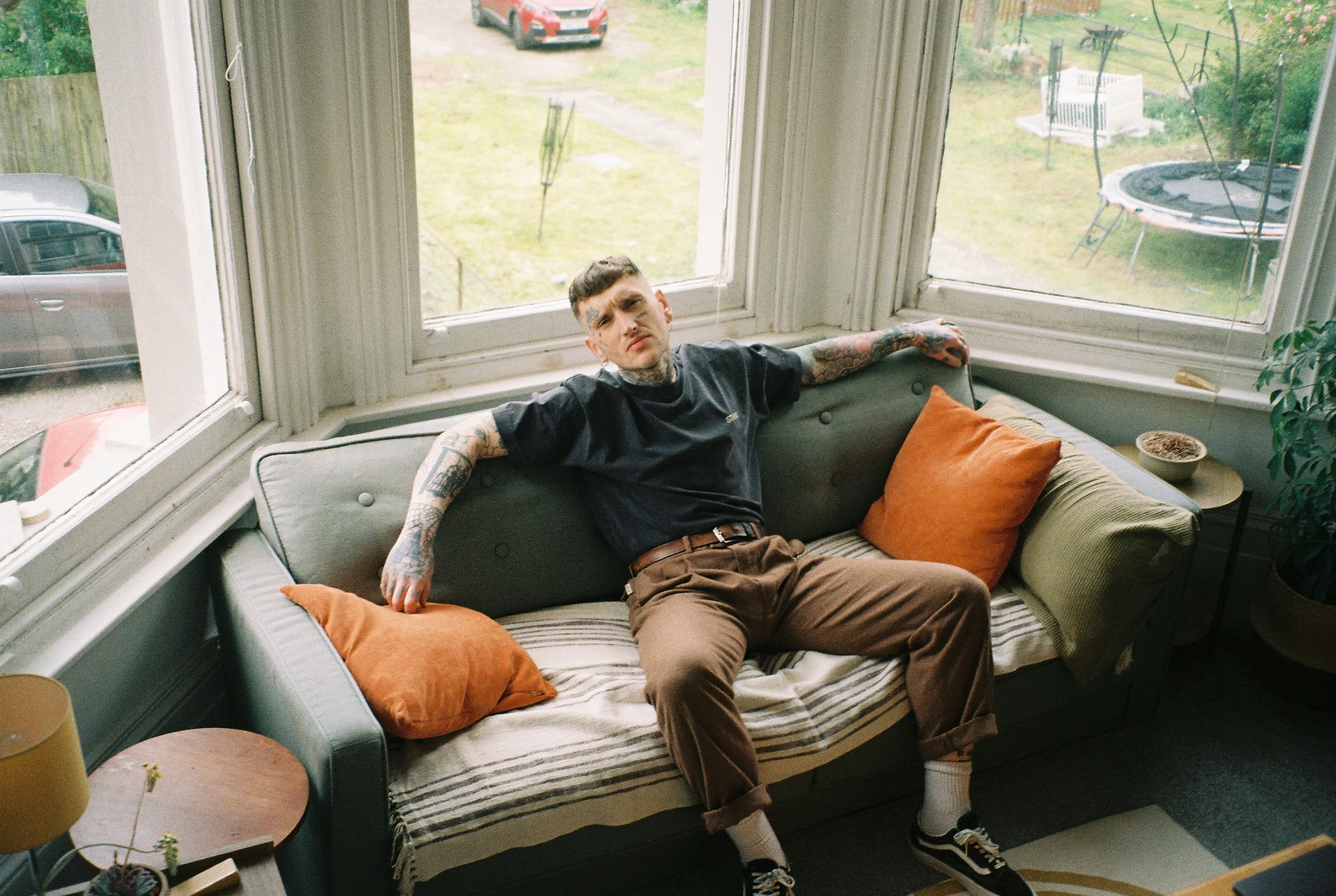 A man with tattoos and a serious expression sitting on a cushioned window seat with orange pillows, with large windows showing a backyard with a trampoline, a car, and outdoor furniture.
