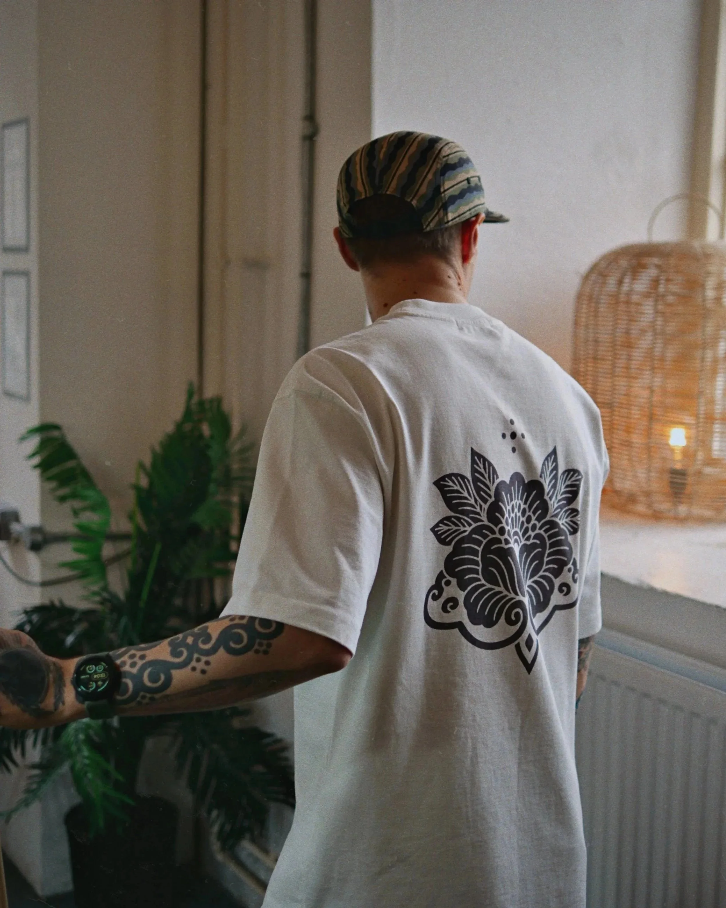 Back of a person wearing a cap and a white t-shirt with a black floral design, standing indoors near a plant and a tall wicker lamp.