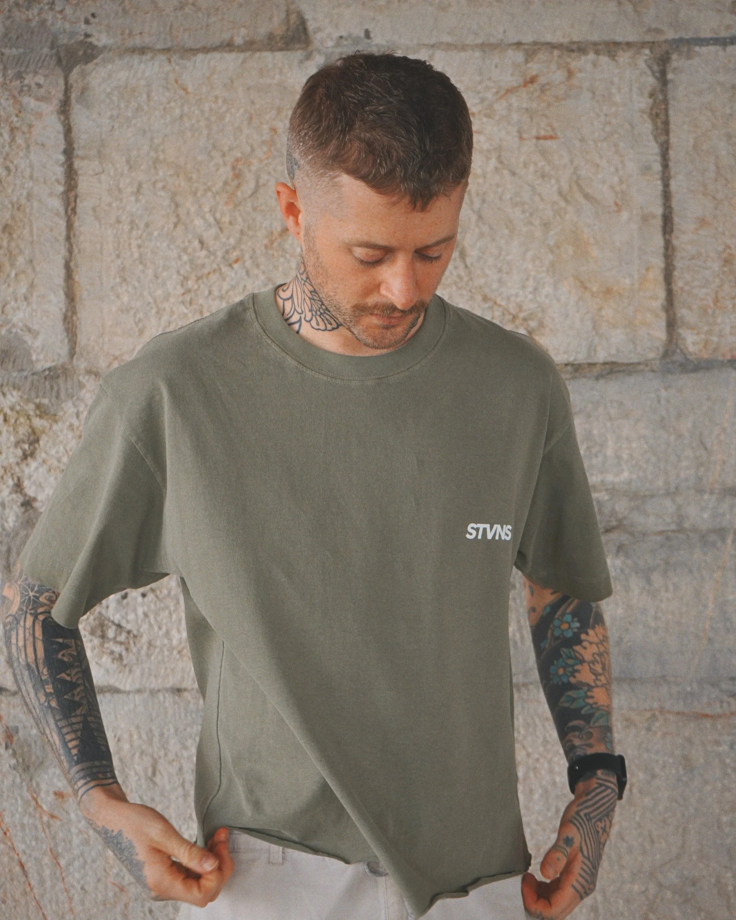 A person with short dark hair, tattoos on their neck and arms, wearing a green T-shirt with "STVNS" written on it, standing indoors against a brick wall.