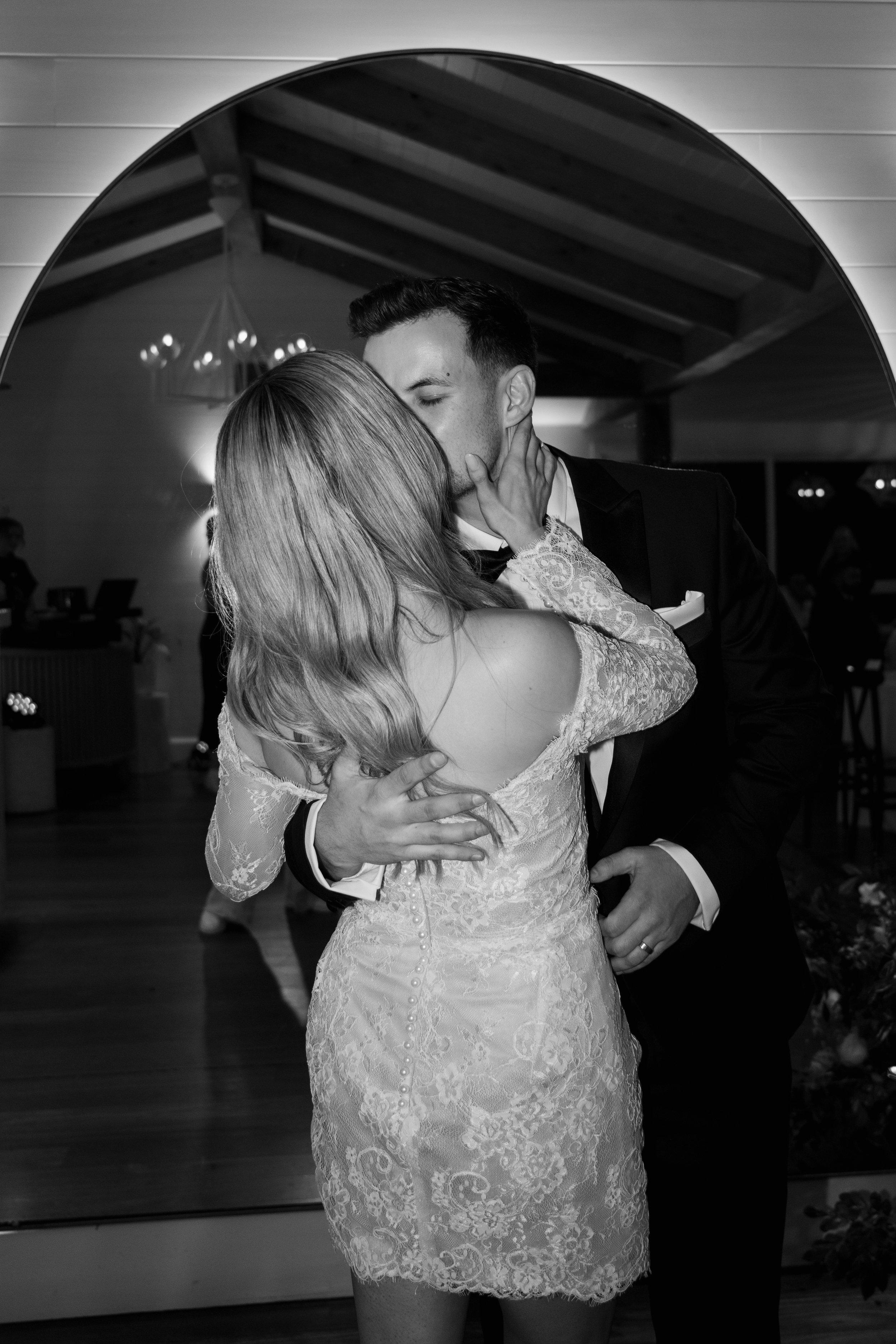 A black-and-white photo of a couple dancing closely indoors, with the woman wearing a lace dress and the man in a tuxedo.