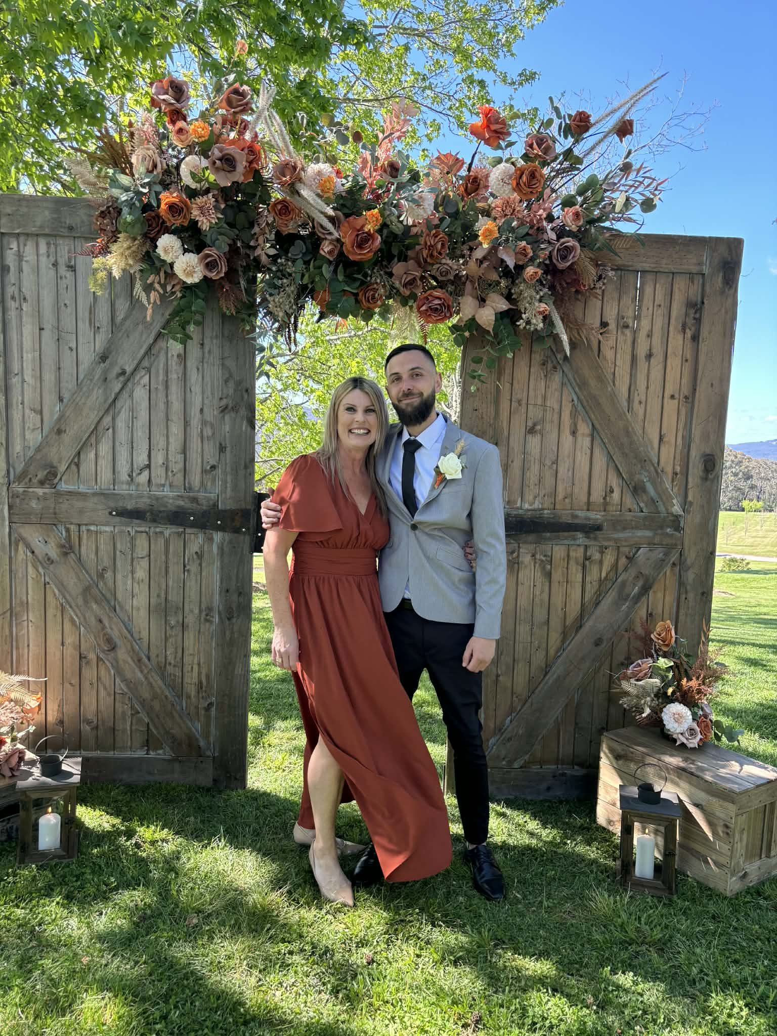 A smiling woman in a rust-colored dress and a smiling man in a light gray suit with a white boutonniere pose together in front of a wooden gate decorated with a large floral arrangement. The setting appears to be outdoors on a sunny day with greenery and a blue sky.