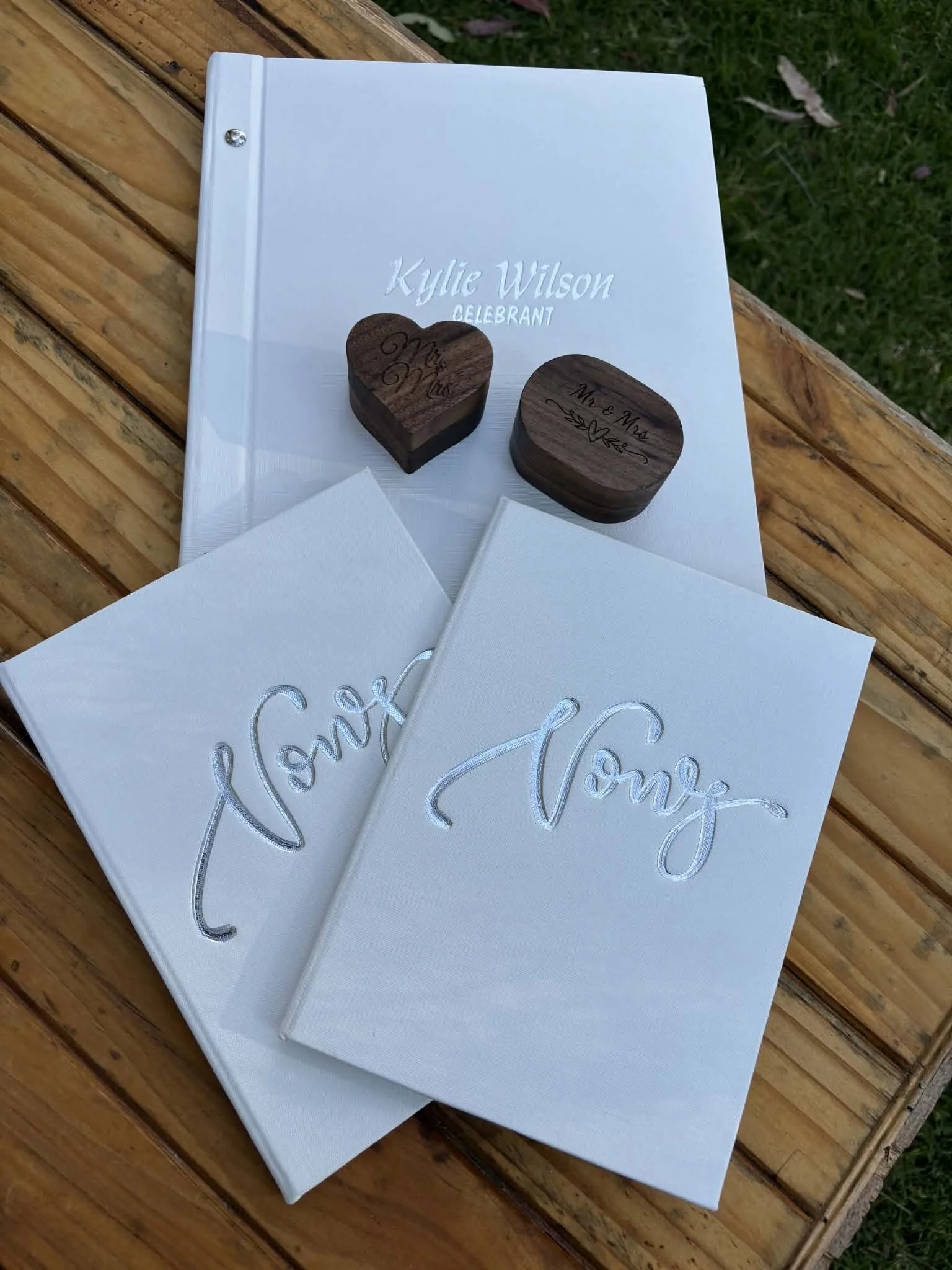 Wedding album with two guest books featuring 'Love' written in script, two wooden engraved boxes labeled 'Mr. & Mrs.', and a white book with 'Kylie Wilson Celebrant' on the cover, all arranged on a wooden surface outdoors.