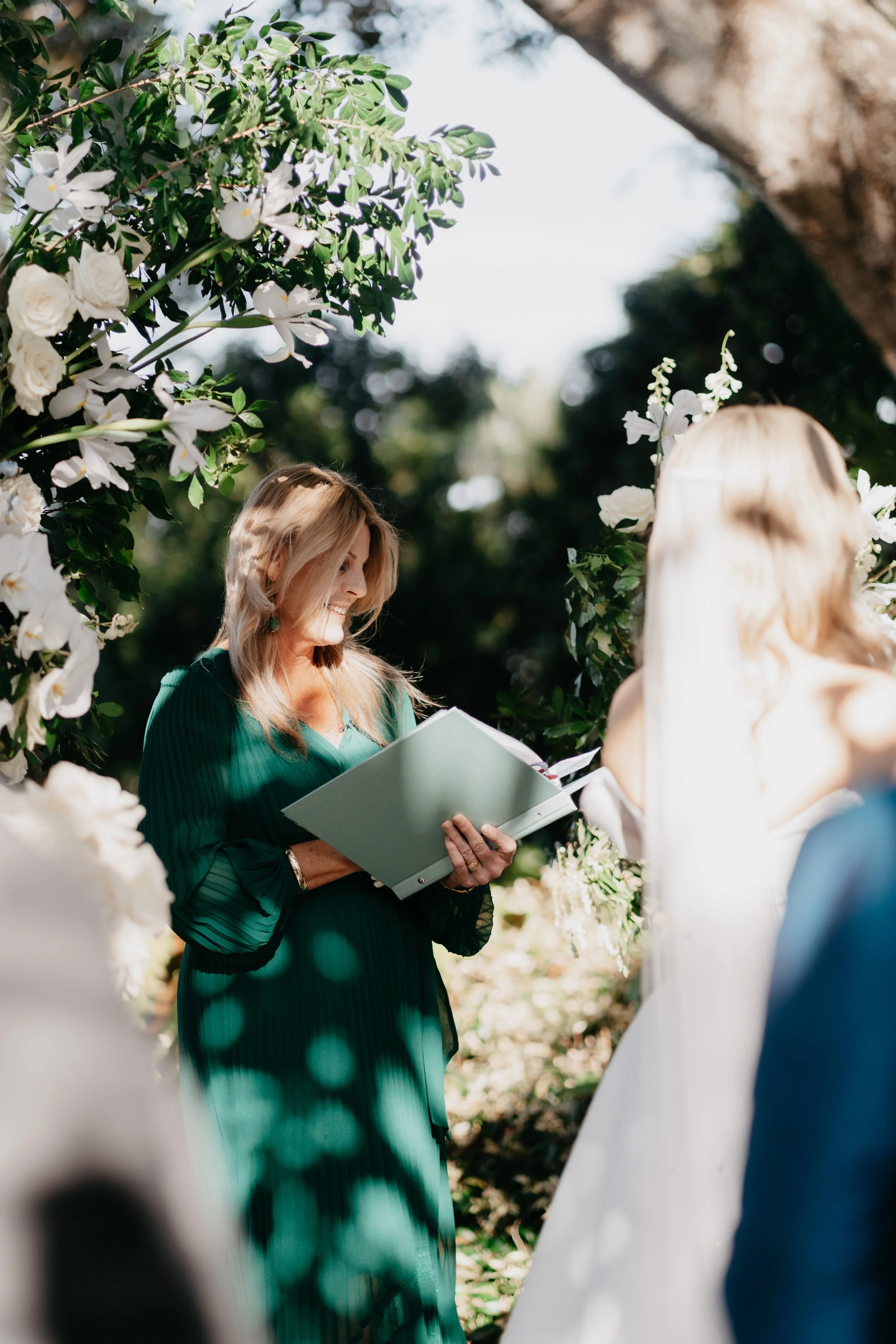 Woman in a green dress officiating a wedding ceremony outdoors, surrounded by white flowers and greenery.