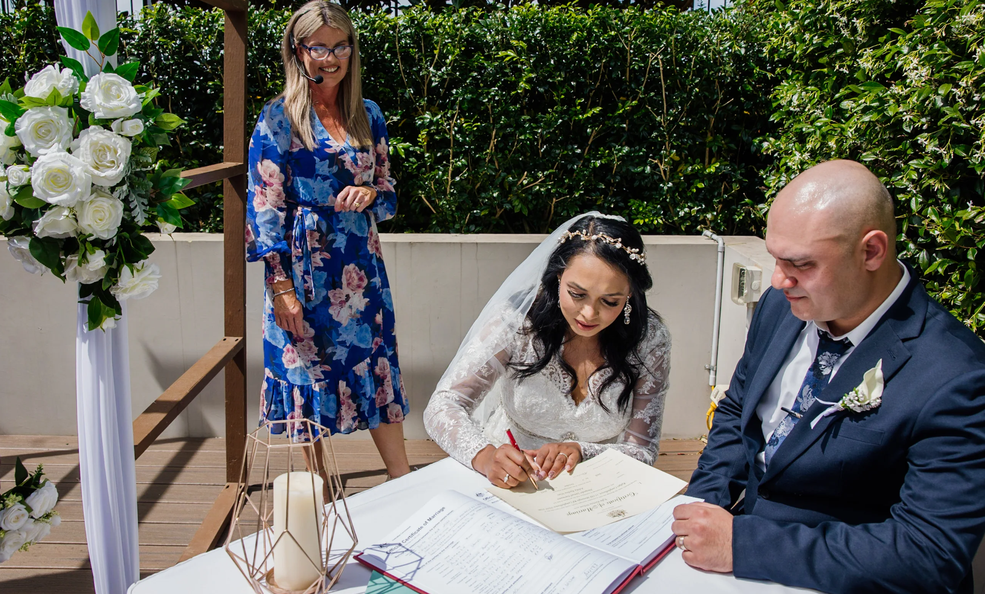 A bride and groom signing marriage documents outdoors with an officiant standing nearby, decorated with white roses and greenery.