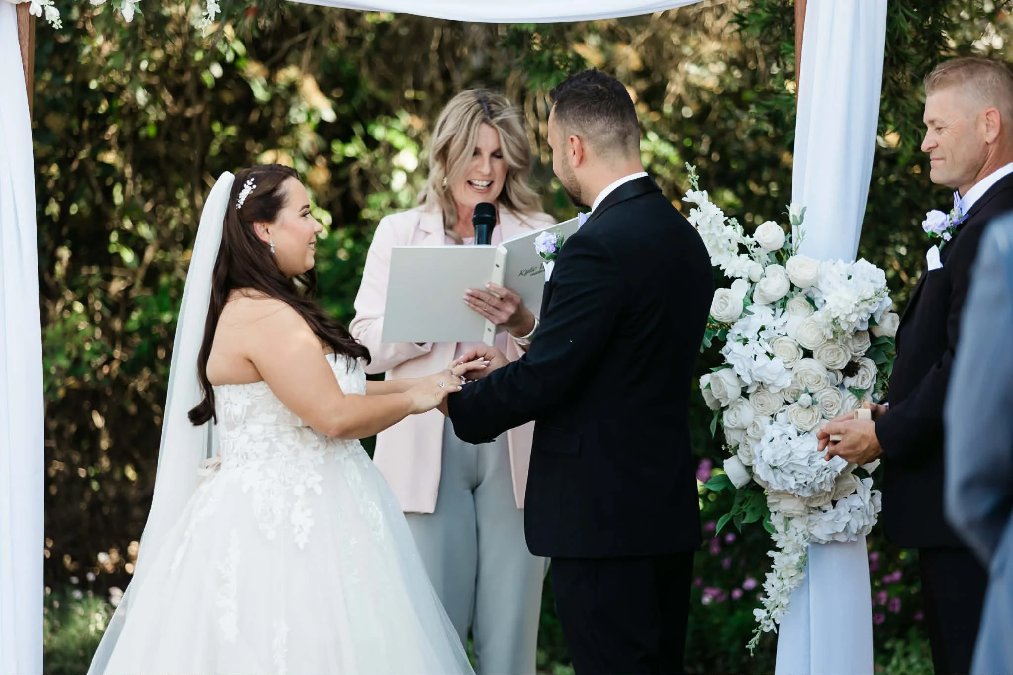 A wedding ceremony outdoors with a bride and groom holding hands, exchanging vows, and an officiant reading from a book. The bride is in a white wedding gown, and the groom is in a black suit. There are large white floral arrangements and a white canopy in the background.
