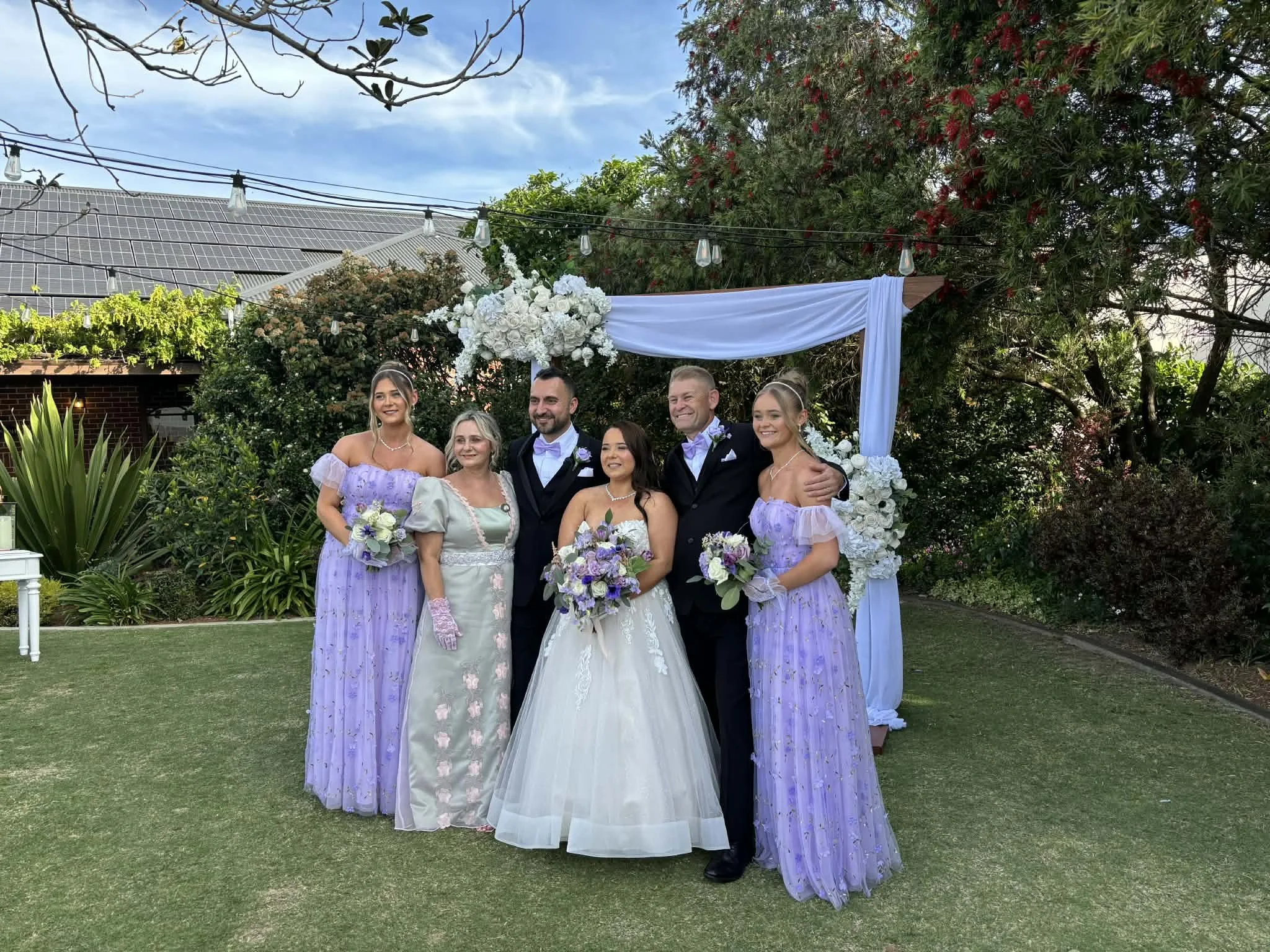A group of six people at a wedding, standing outdoors in front of a decorated arch with white flowers and fabric, surrounded by greenery. The bride is in a white wedding gown, and the groom and two women are in black tuxedos with purple dresses.