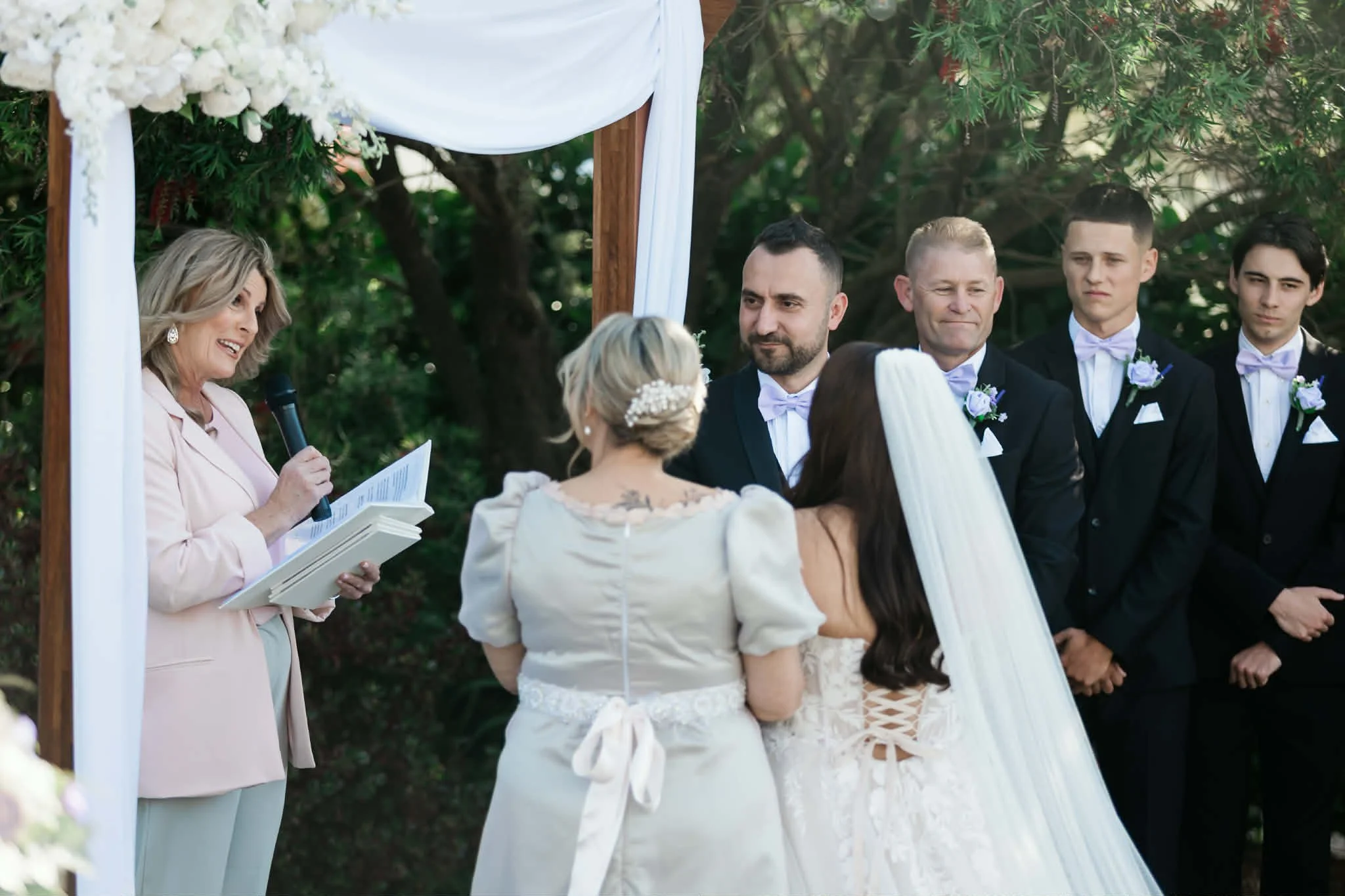Couple exchanges vows during outdoor wedding ceremony under a white fabric arch, surrounded by trees, with officiant reading from a book, and bridesmaids and groomsmen standing nearby.